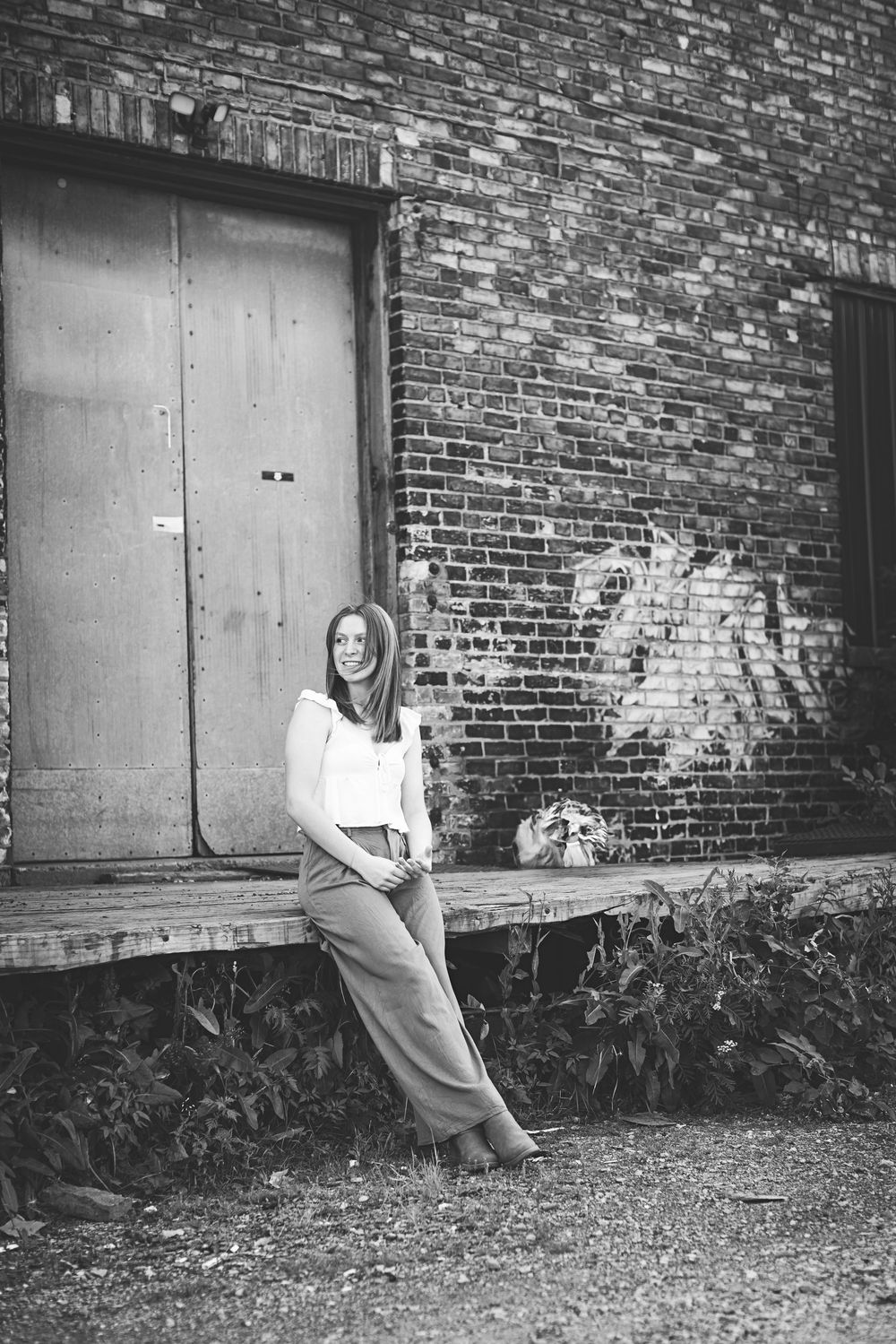 Black and white photo of a casual pose against a brick wall with metal door near an industrial building exterior.