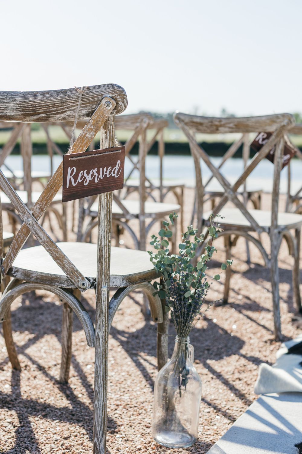 A row of rustic wooden chairs with reserved signs and small floral arrangements decorate an outdoor wedding ceremony setup.