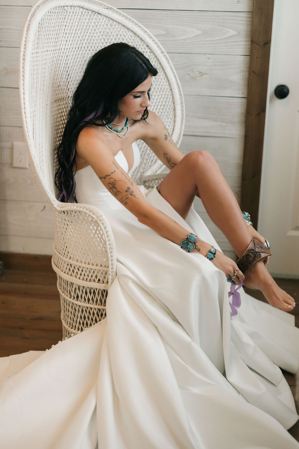 A person in a white wedding dress sits in a vintage wicker peacock chair against light wood paneling.