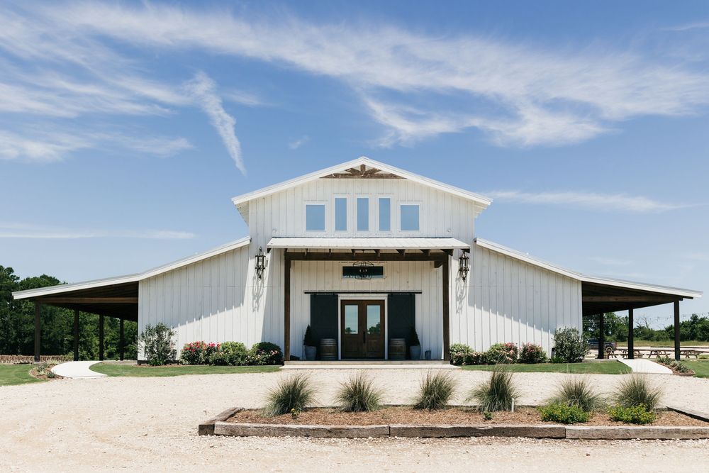 Modern white barn-style wedding venue with large glass windows and covered porches on a sunny day.