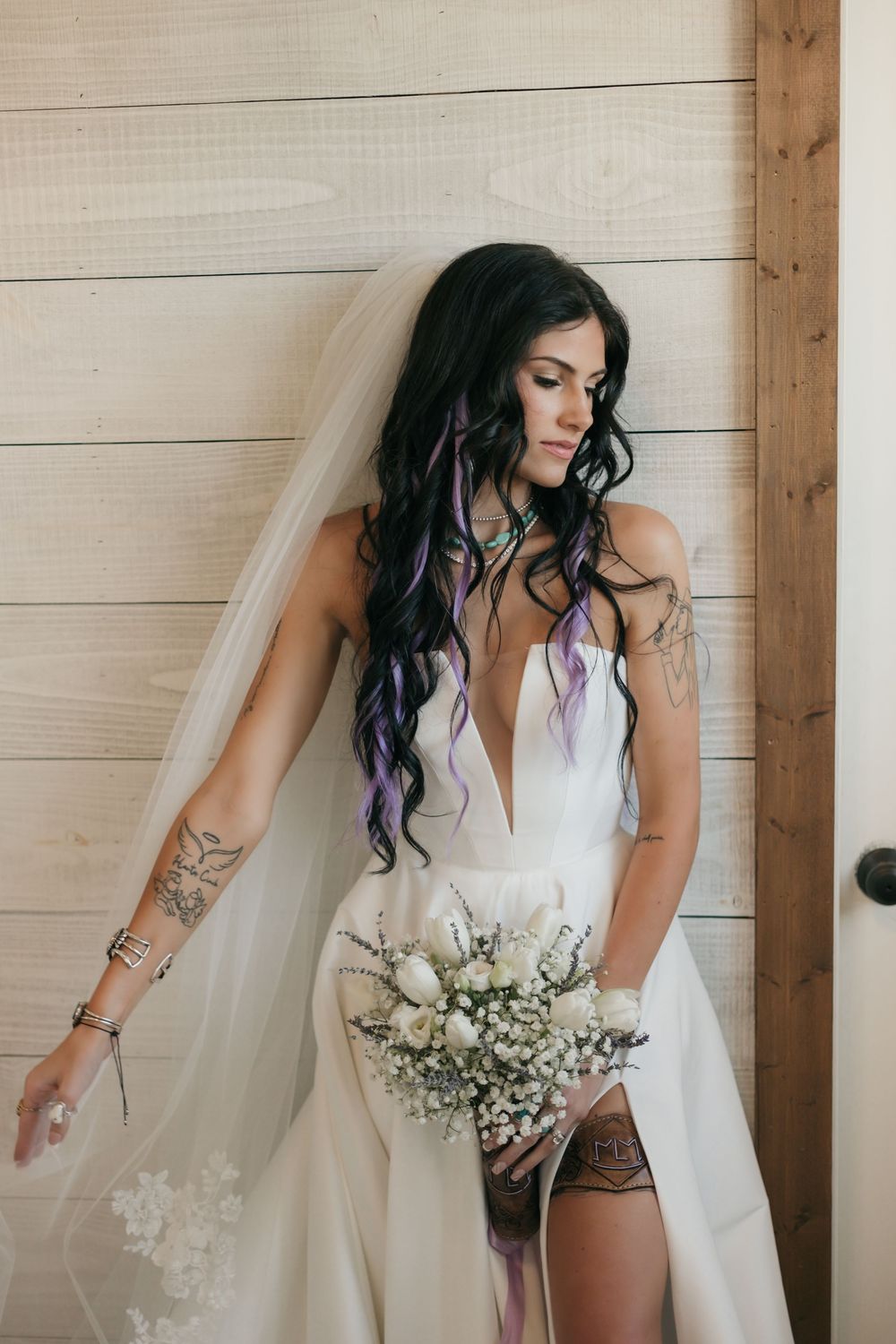 Bride in white dress with purple highlights in hair holds delicate white bouquet against wooden wall background.
