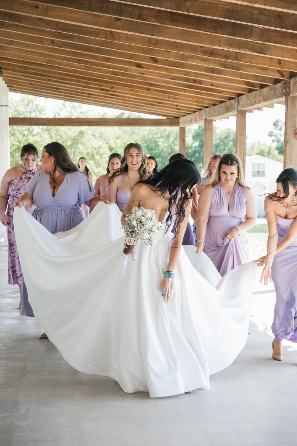 A group of bridesmaids in lavender dresses holding up the flowing train of a white wedding dress in a covered outdoor venue.