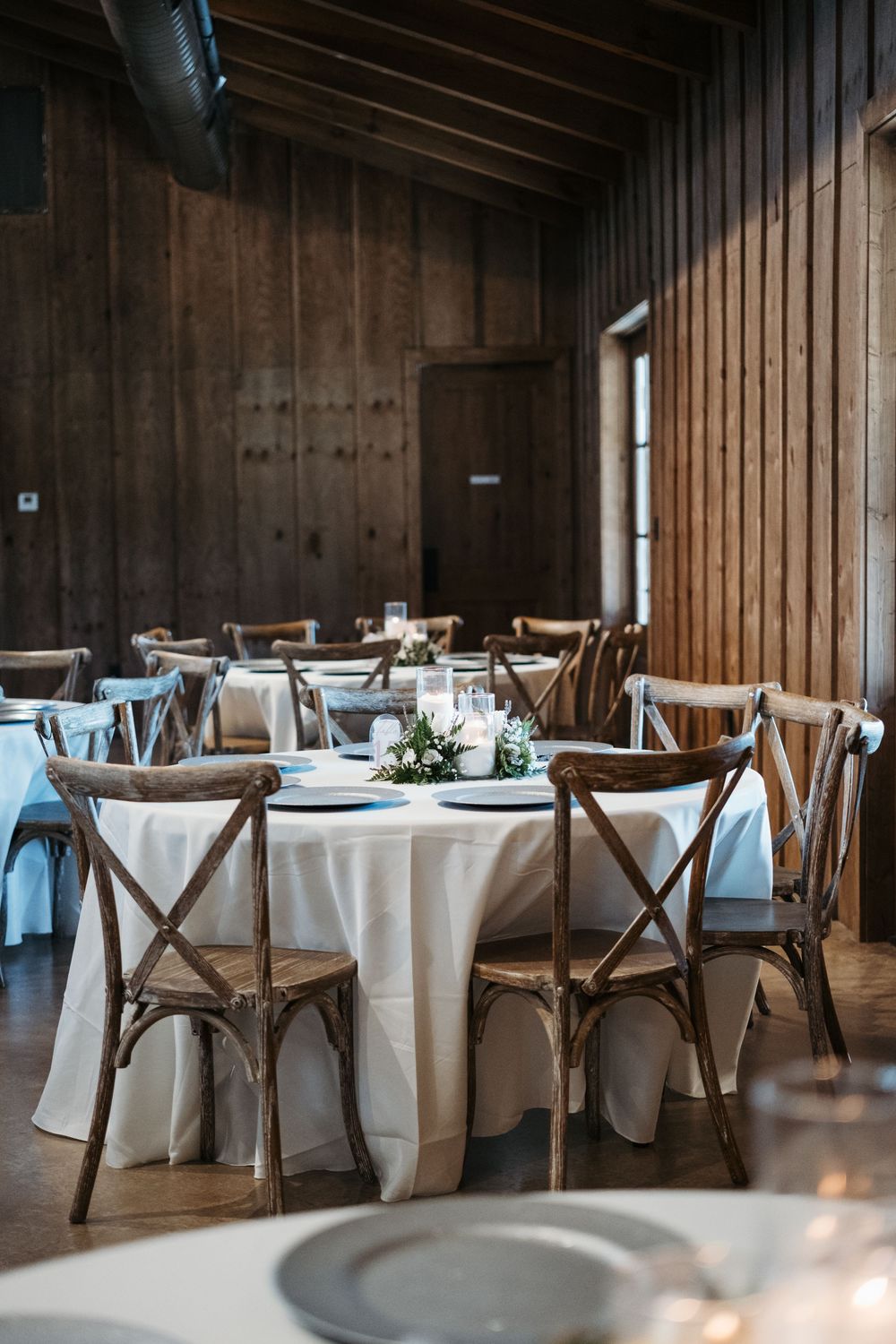 A rustic wedding reception venue with wooden walls, white tablecloths and cross-back chairs in warm natural lighting.