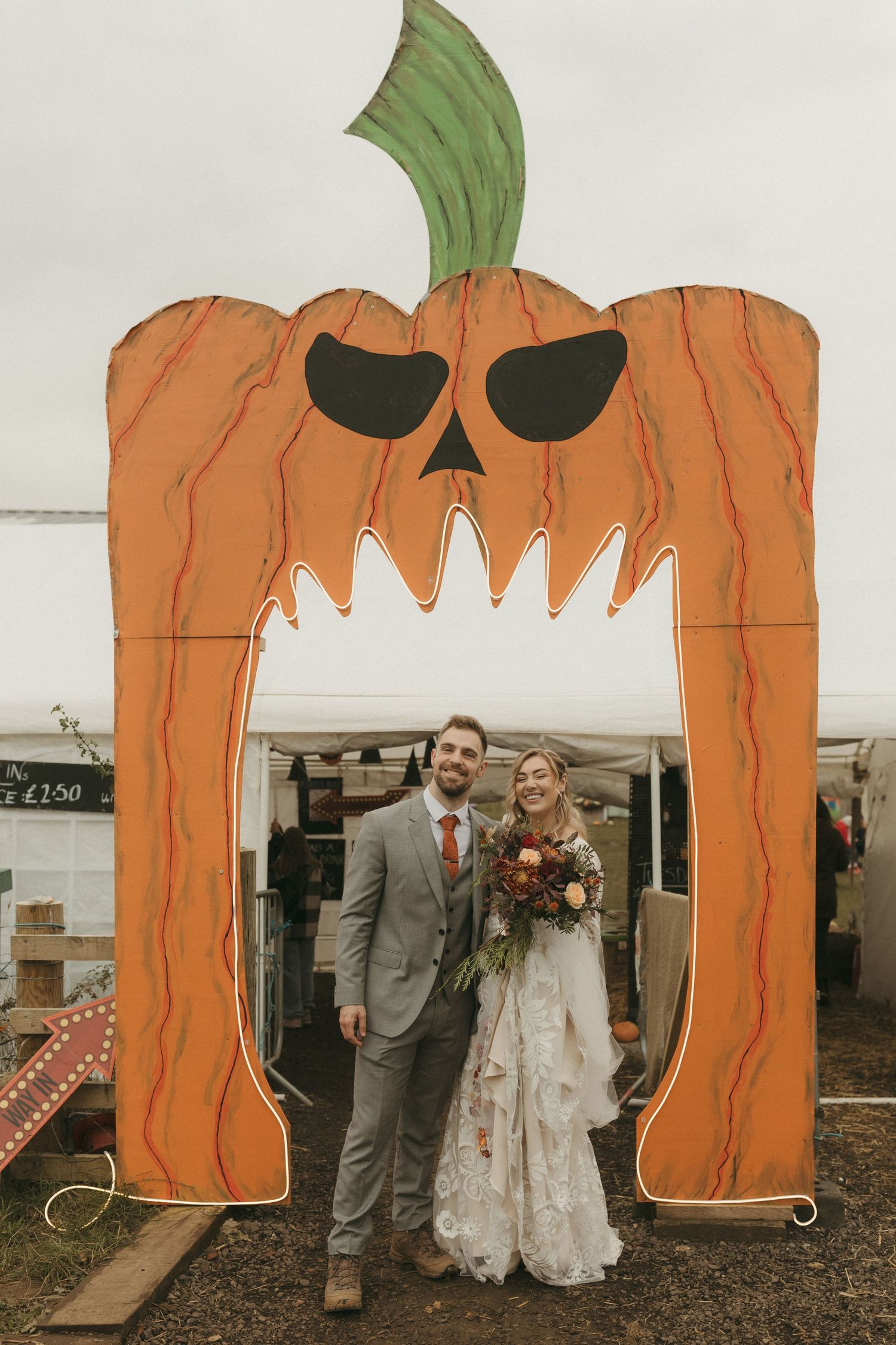 A couple poses under a large orange pumpkin-shaped arch with a spooky face during their Halloween-themed wedding.