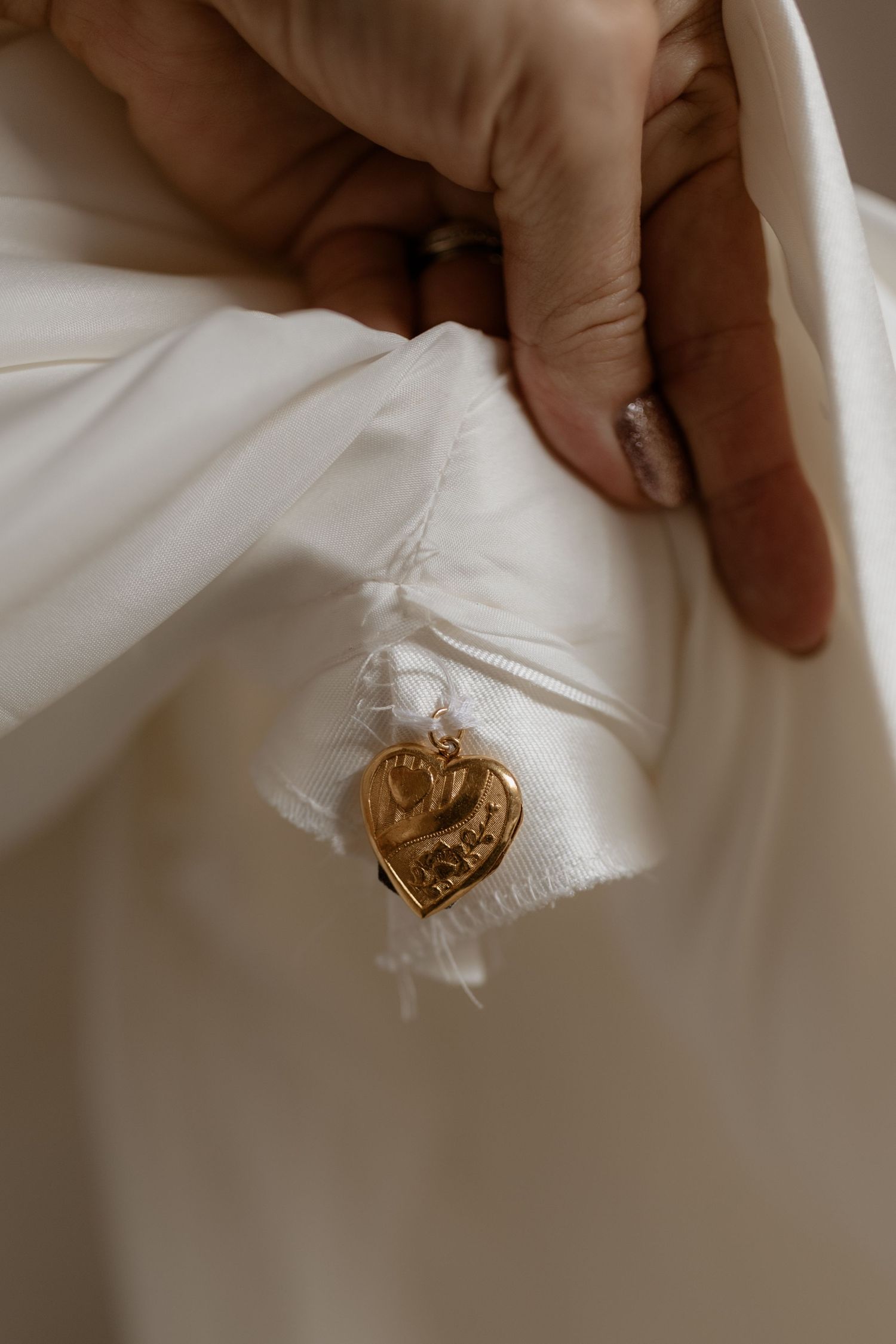 A golden heart-shaped locket or charm detail shown against white fabric, with fingers delicately touching the material.