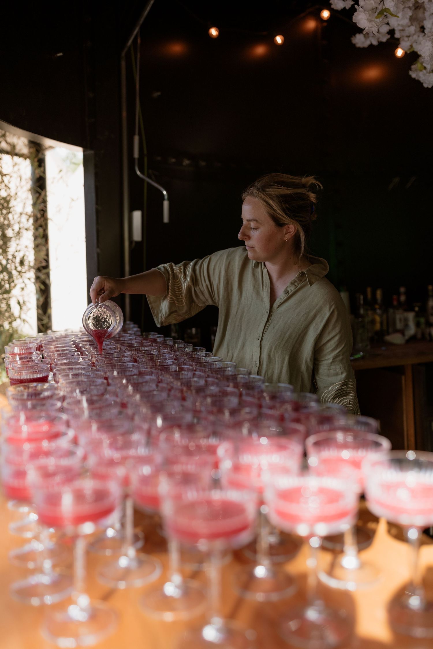 Someone pours drinks into multiple coupe glasses arranged on a wooden bar counter with warm lighting in the background.
