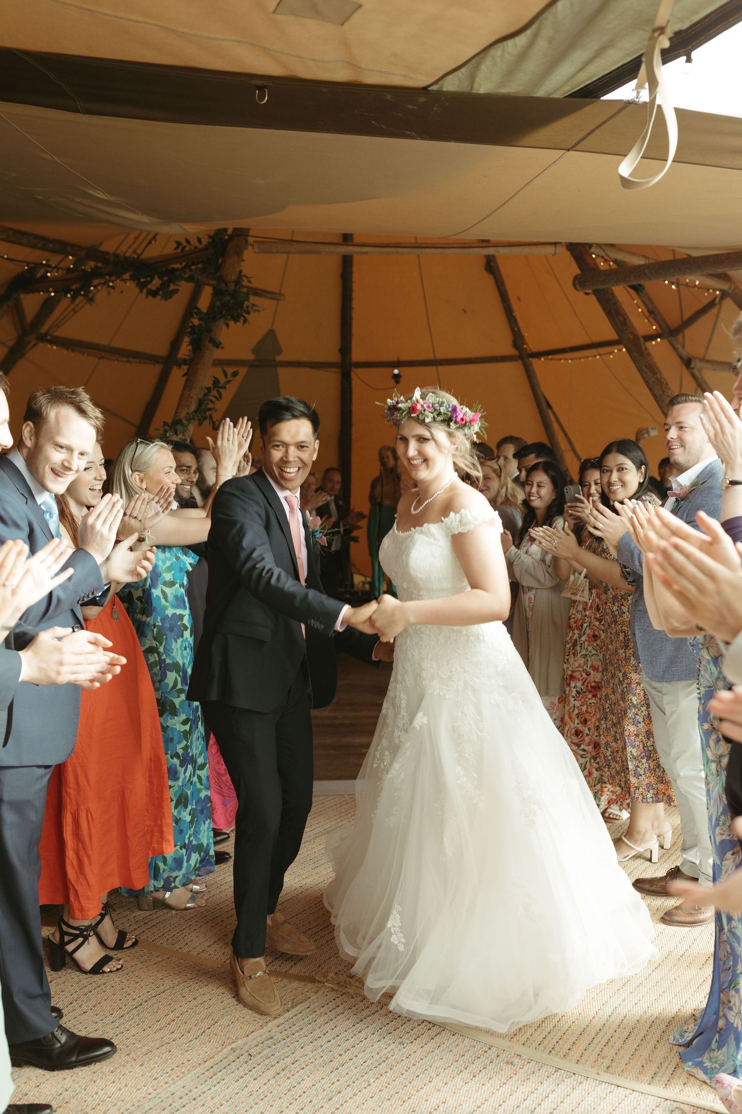Wedding celebration under a teepee tent with guests applauding as couple holds hands on rustic carpeted floor.
