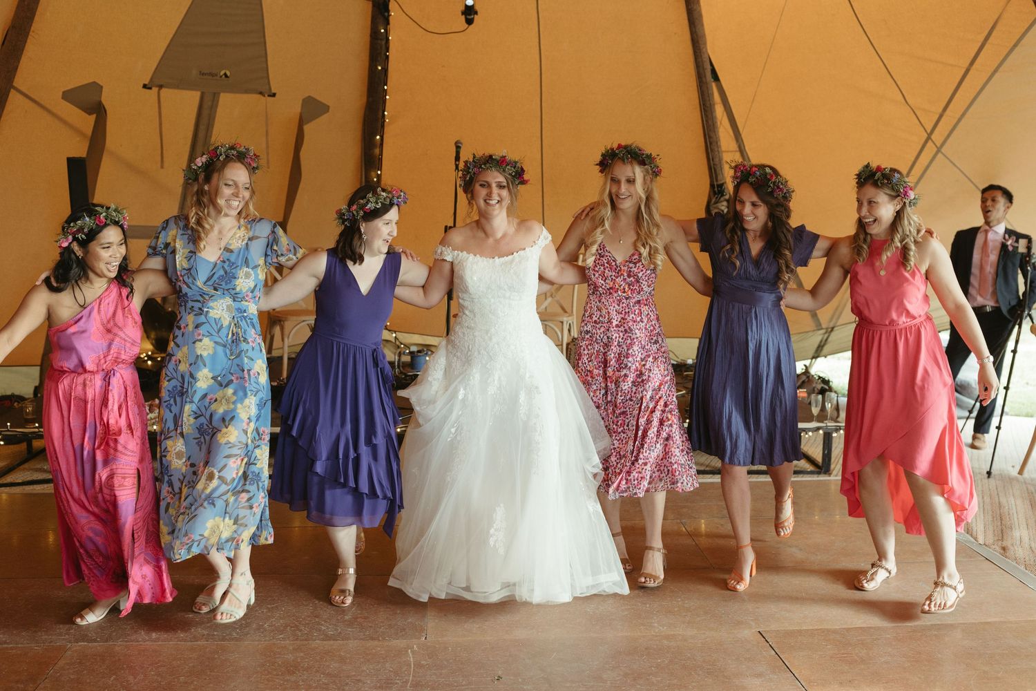 A group of wedding guests in colorful dresses dance together under a large tent during a celebration.