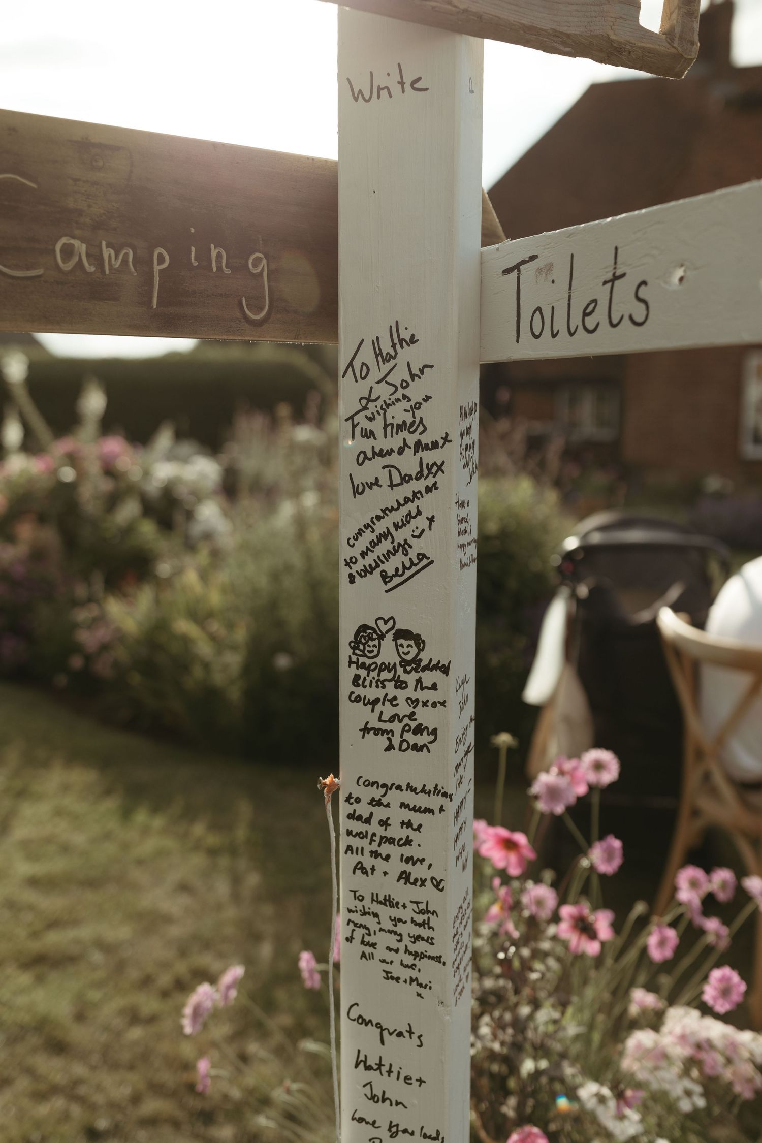 A white support beam with handwritten notes and messages, surrounded by pink flowers in a camping area.