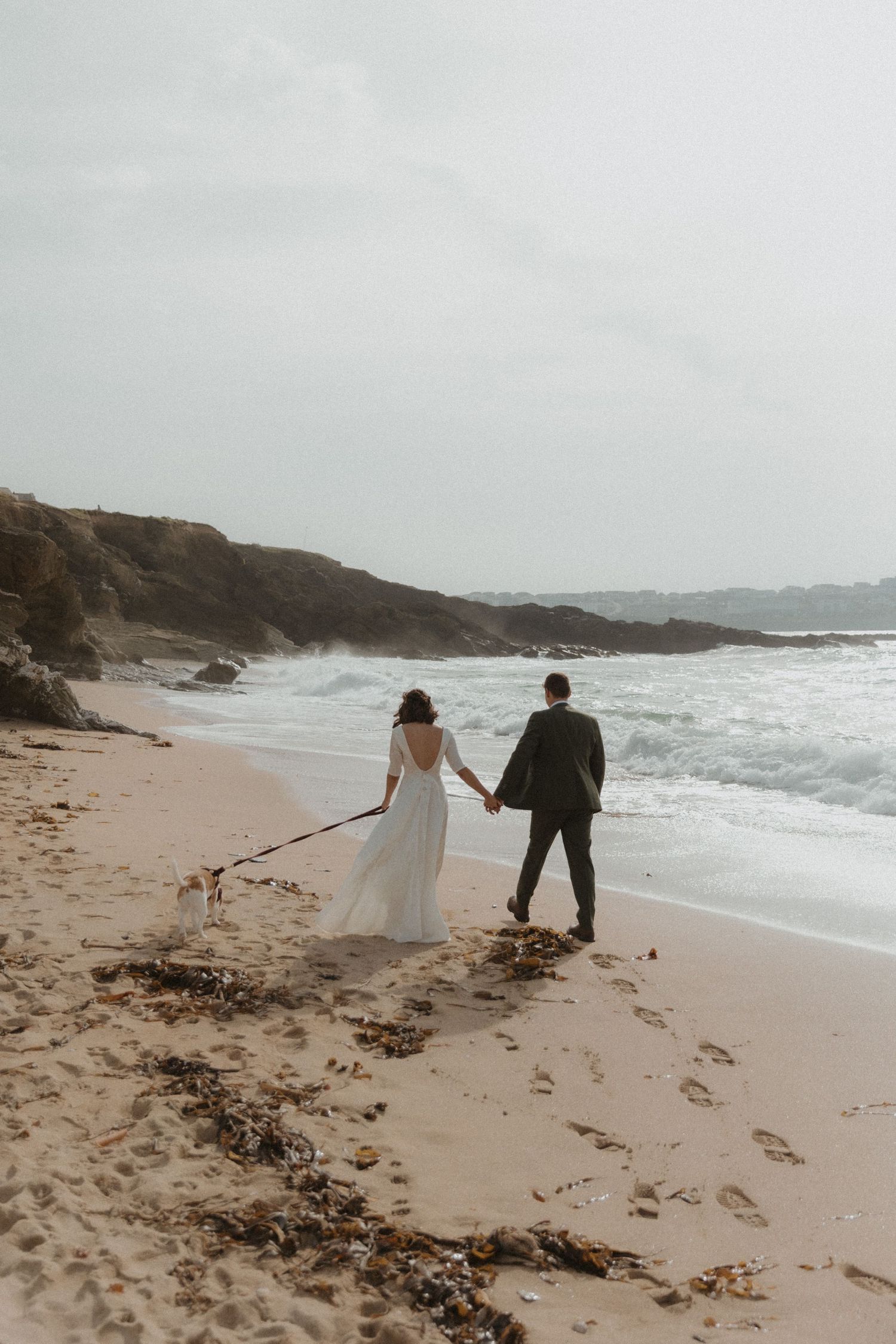 A couple in wedding attire walks hand in hand along a misty beach with dramatic waves crashing against rocky cliffs.