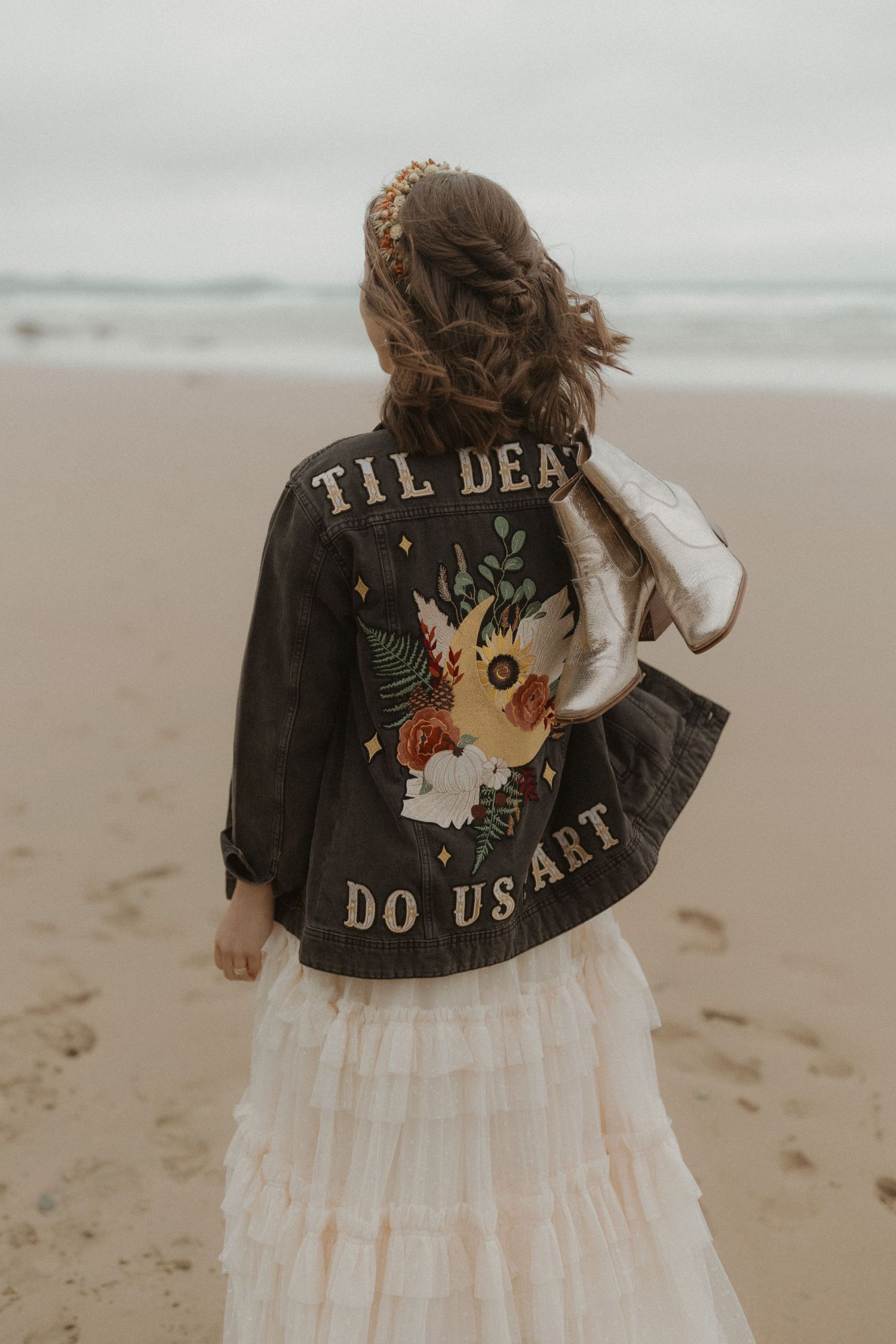 Side view of leather jacket with embroidered "TIL DEATH DO US PART" text on back, paired with flowy white dress.