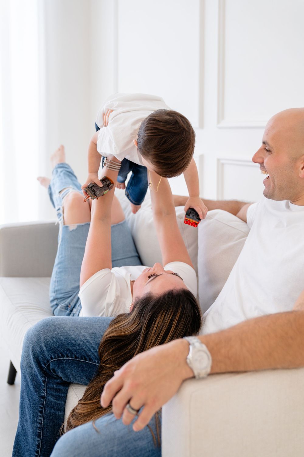 A happy family plays together on a white couch, lifting a young child up high with joyful expressions.