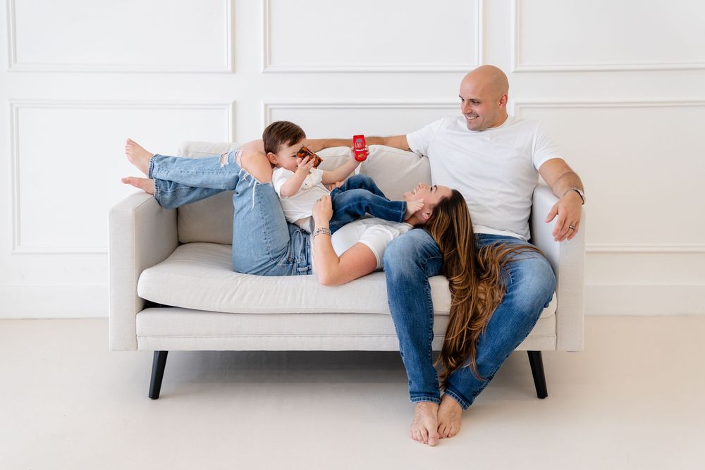 A father and children playfully relax together on a light gray modern couch in a bright minimalist room.