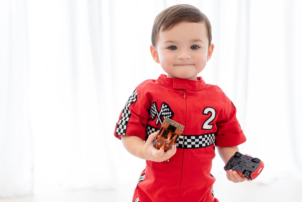Child wearing red racing-themed outfit holds toy cars against white backdrop.