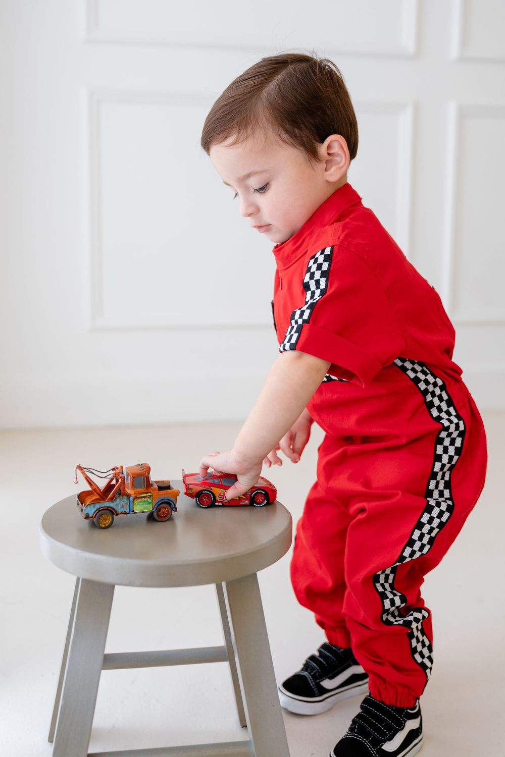 A child in a red racing jumpsuit with checkered stripes plays with toy cars on a small gray table.