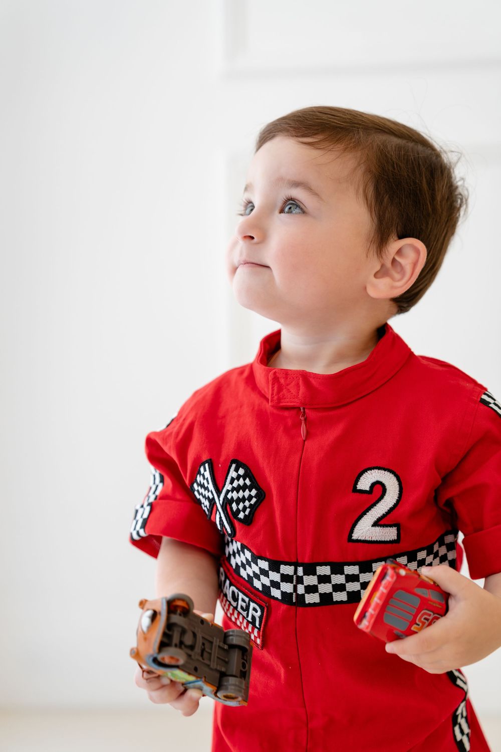 Child wearing red racing jumpsuit with number 2 plays with toy cars against white background.