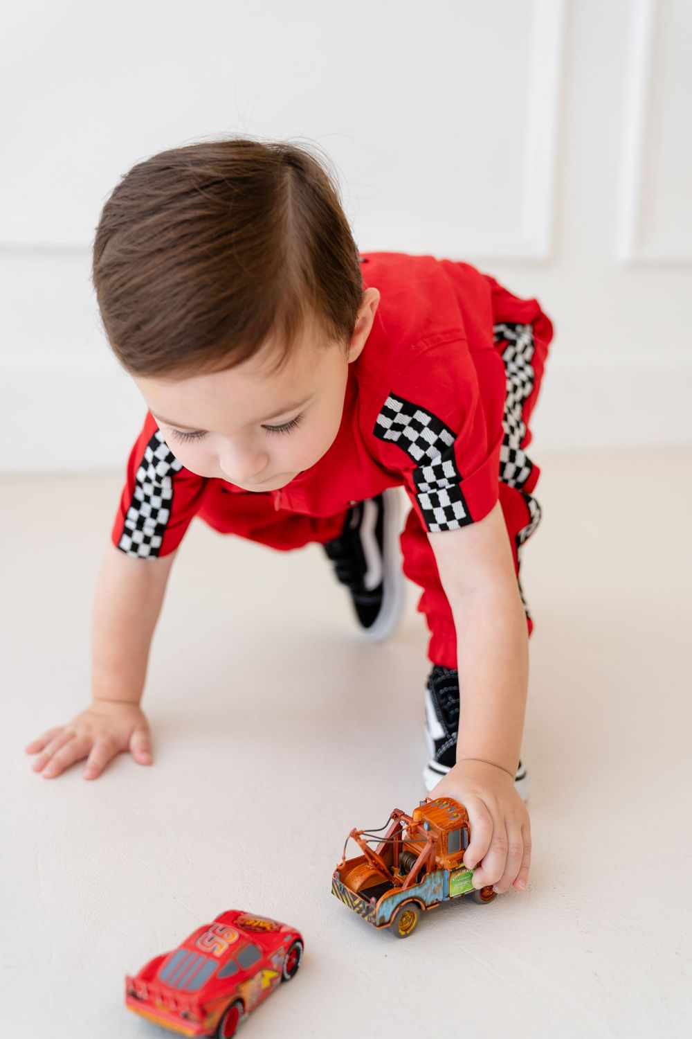 Child in red racing outfit reaches down to play with toy cars on white surface.
