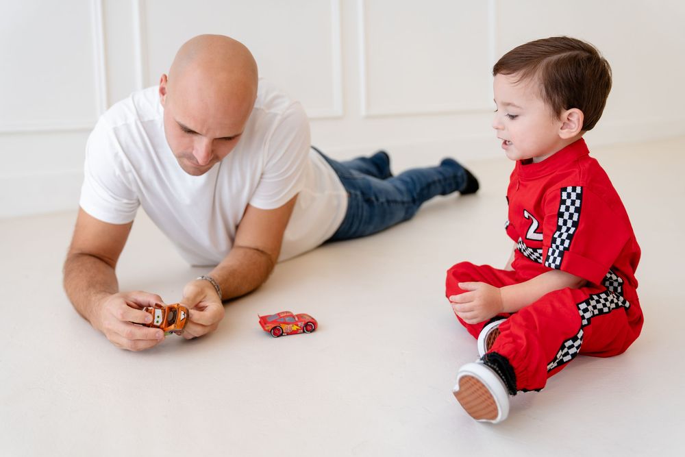 A child in red racing pajamas plays with toy cars on a white floor while an adult lays nearby.