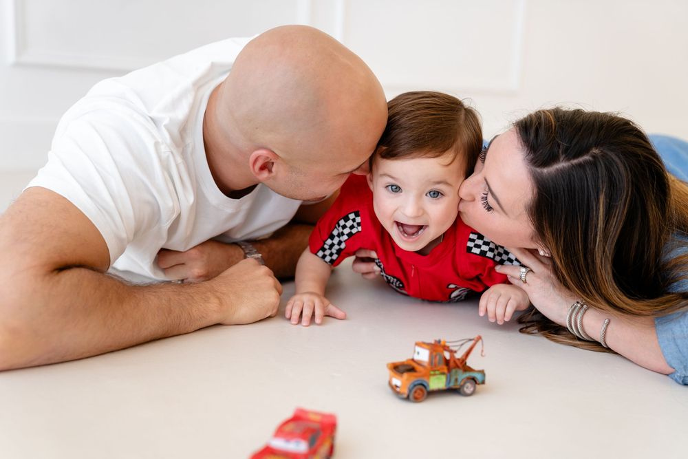 A happy family lies on the floor playing with toy cars together on a white surface.