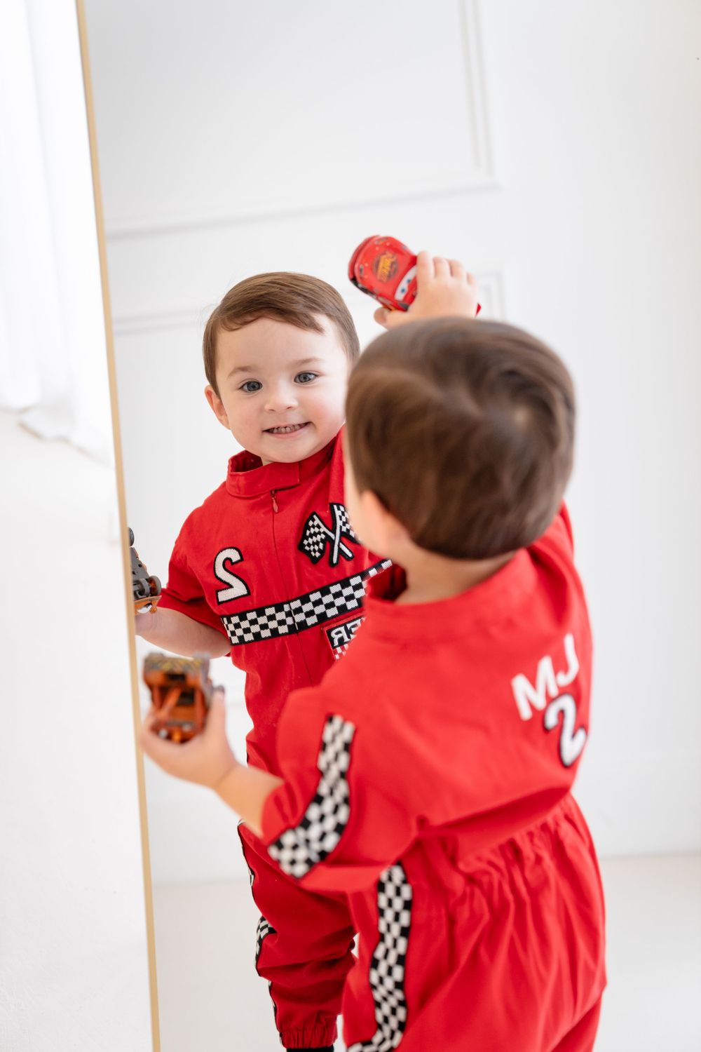 A young toddler in a red racing outfit looks at their reflection in a mirror while holding a toy.