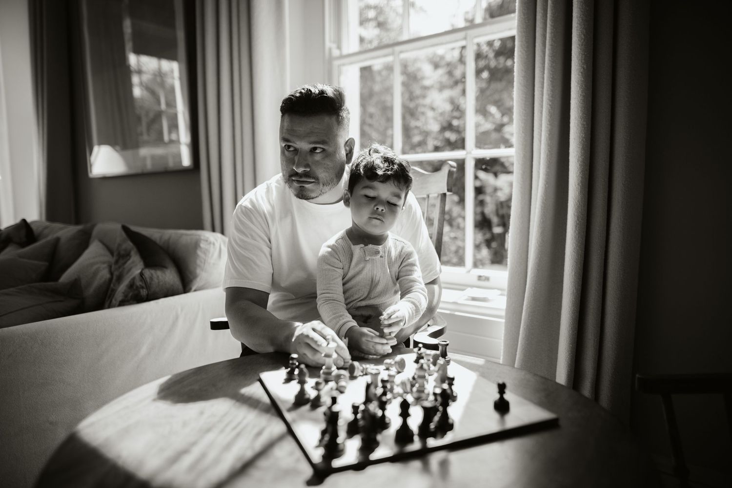 Chess pieces are arranged in gameplay with a player in contemplative thought beside a sunlit window.