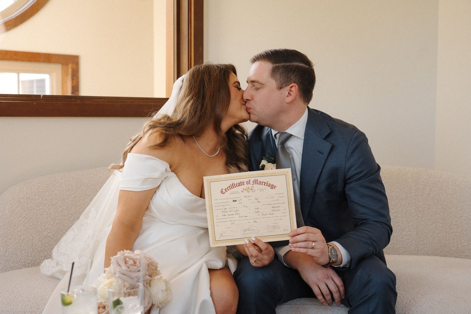 A couple in wedding attire share an intimate moment while holding a marriage certificate on a beige couch.