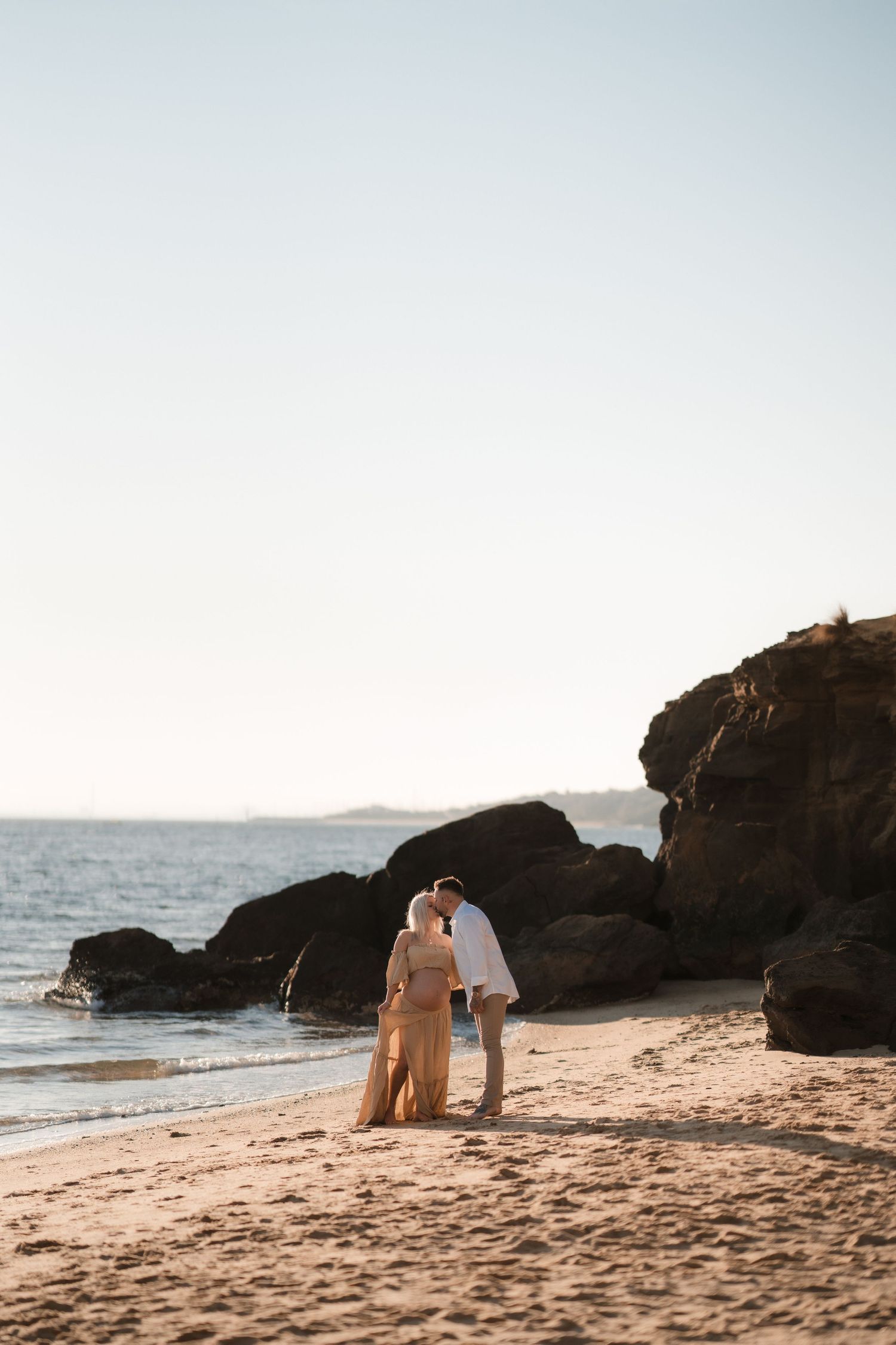 A couple walks along a scenic rocky beach at sunset with waves crashing against the coastline.