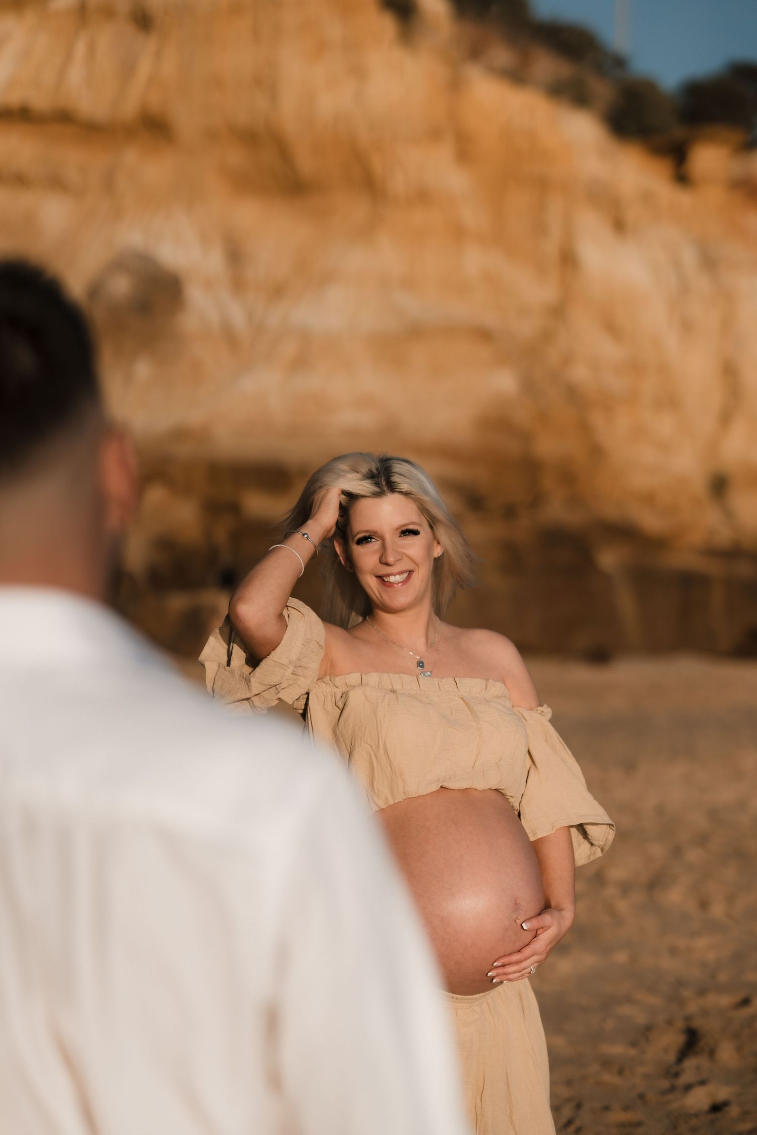 A maternity photoshoot at sunset features a flowing beige off-shoulder dress against dramatic coastal cliffs.