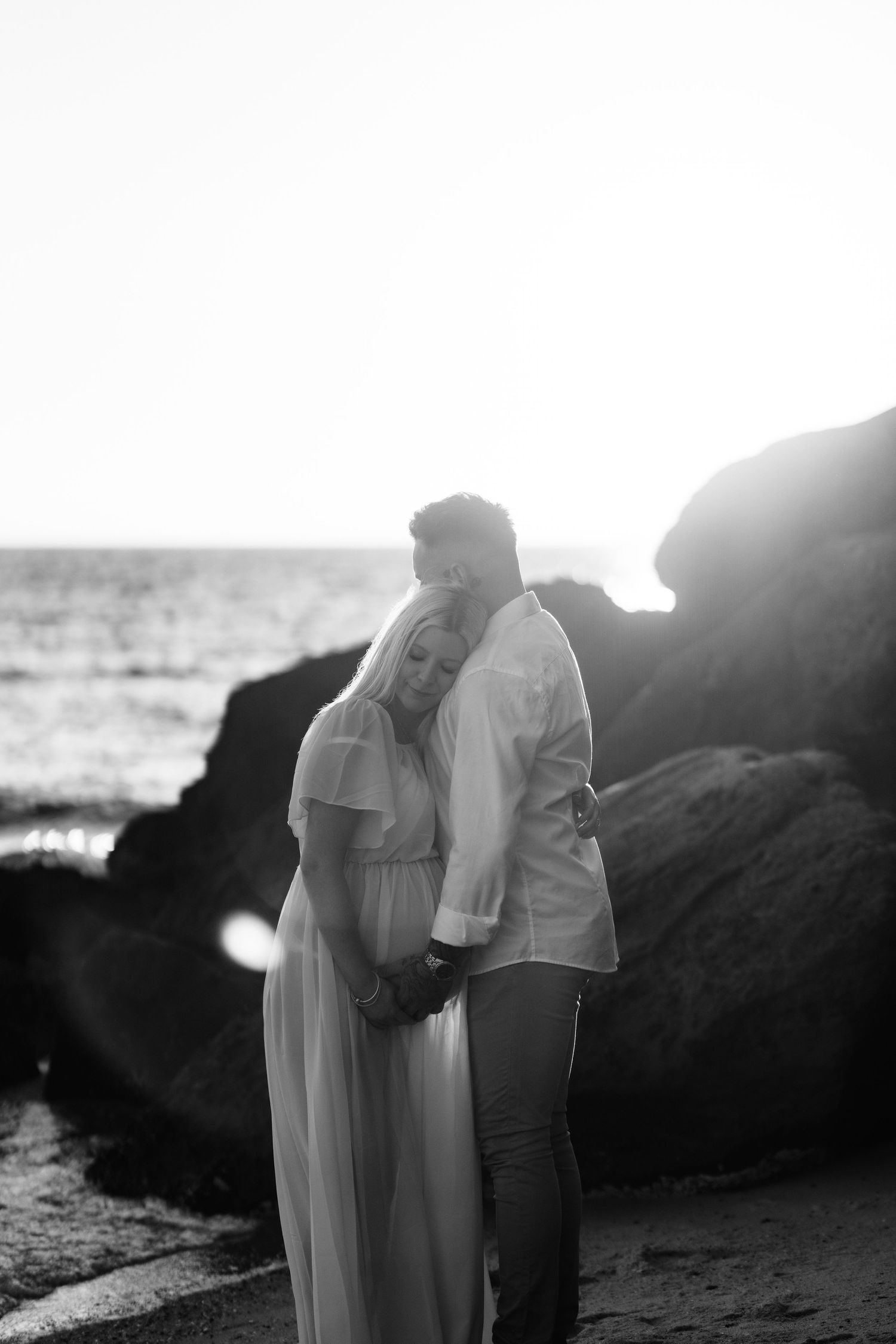 A romantic black and white composition of a couple standing close together on a rocky shoreline at sunset.