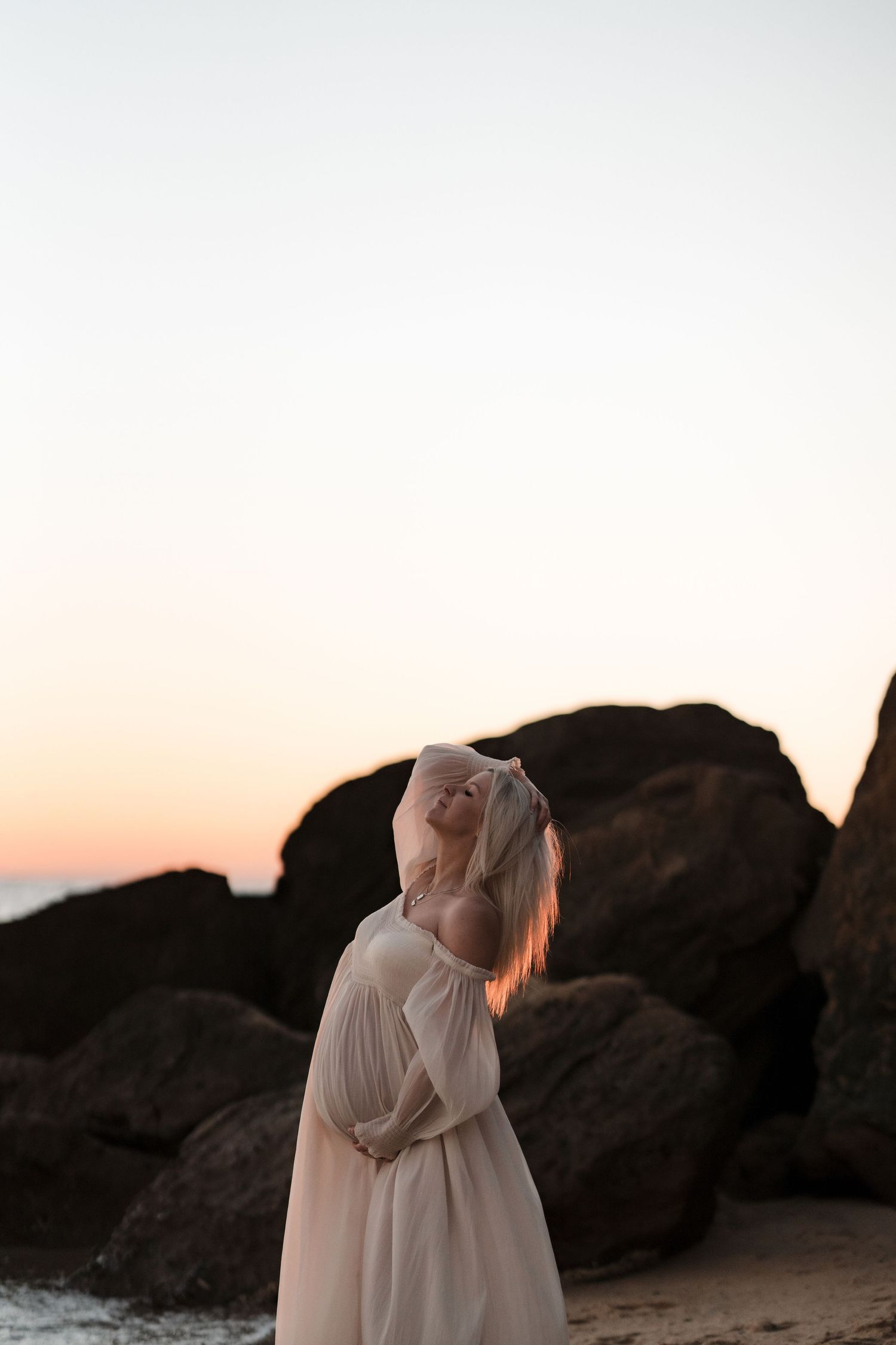 A maternity photo at sunset shows a flowing cream dress silhouetted against large coastal rocks.
