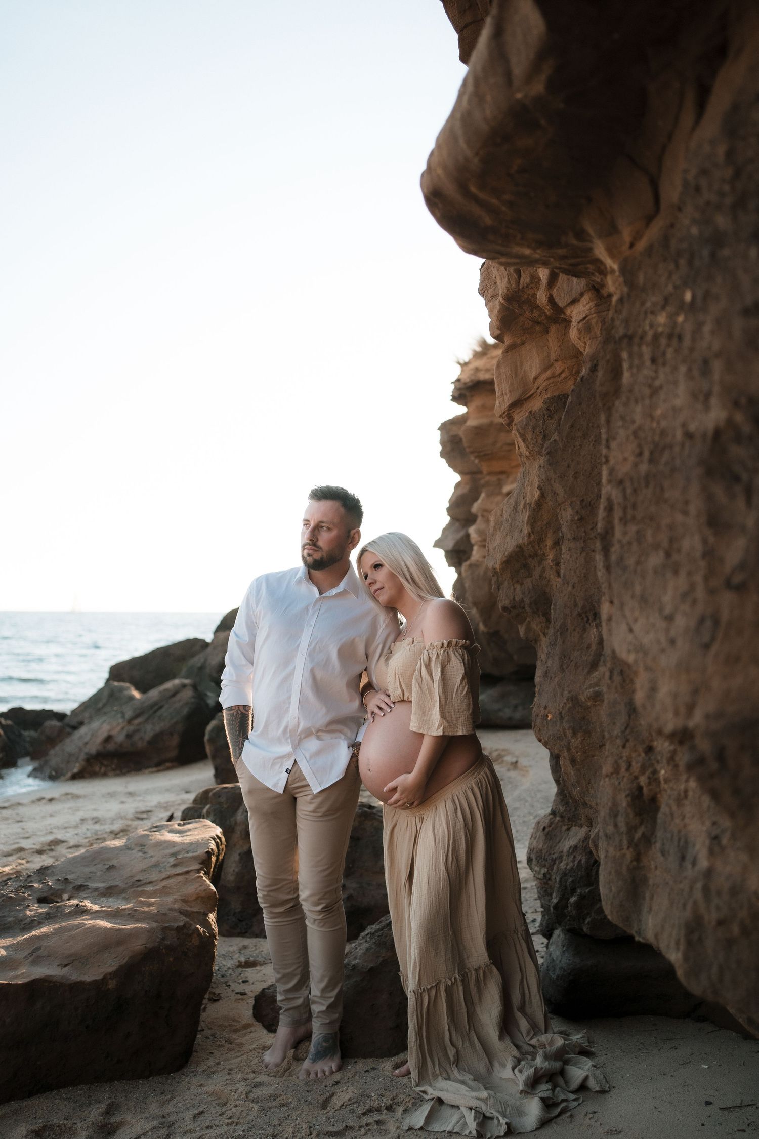 A couple stands together on a rocky beach at sunset, posing beneath a coastal cave formation by the ocean.