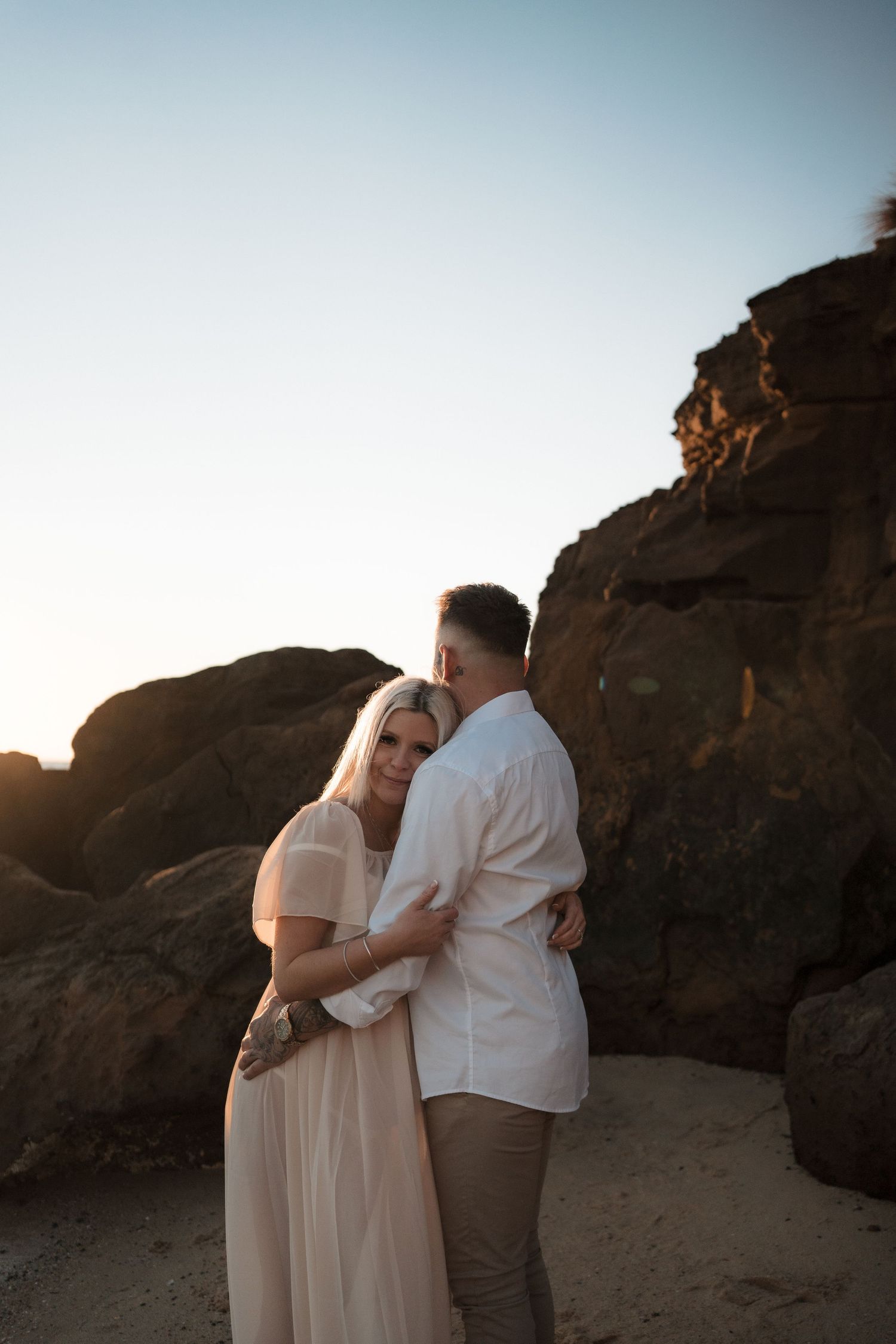 An intimate maternity portrait captures a tender embrace against rocky coastal cliffs at sunset.