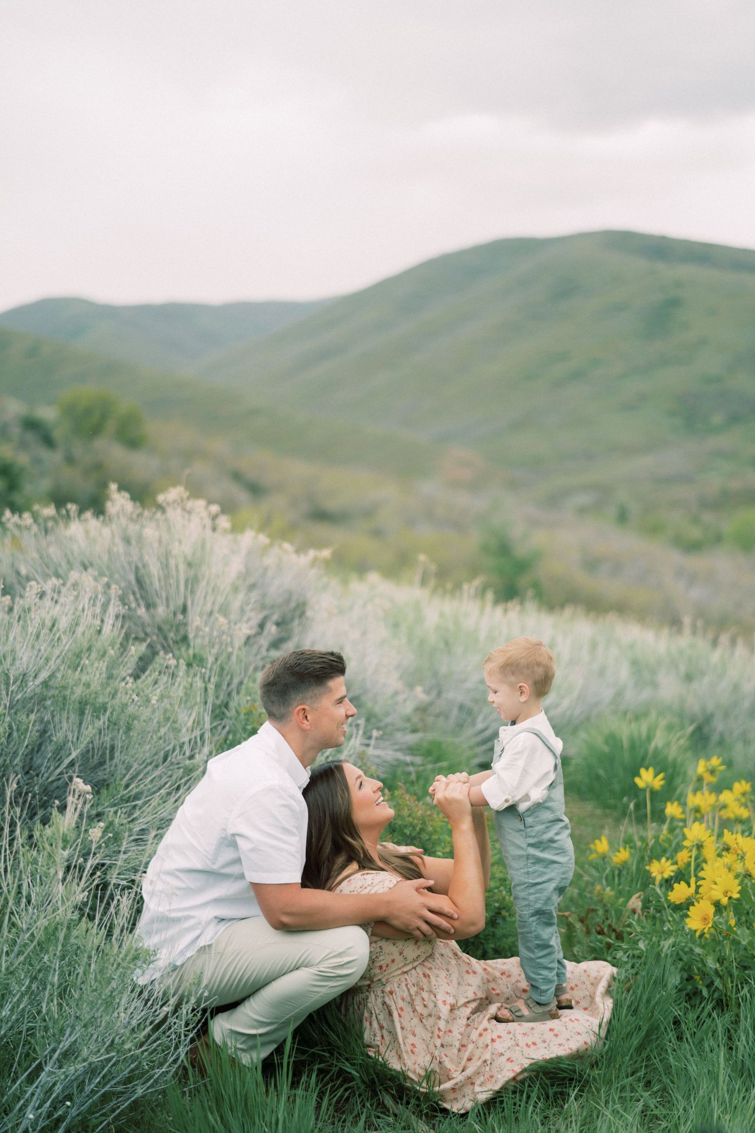 Family lounges together in wild sage brush field with yellow wildflowers and rolling green hills in background.