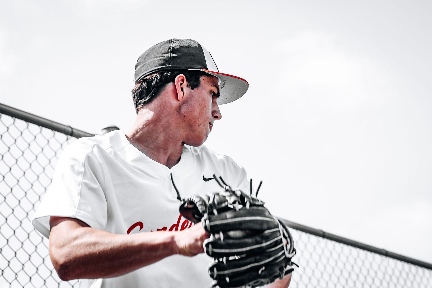 Baseball player in white uniform and cap adjusts his glove while standing next to chain link fence.