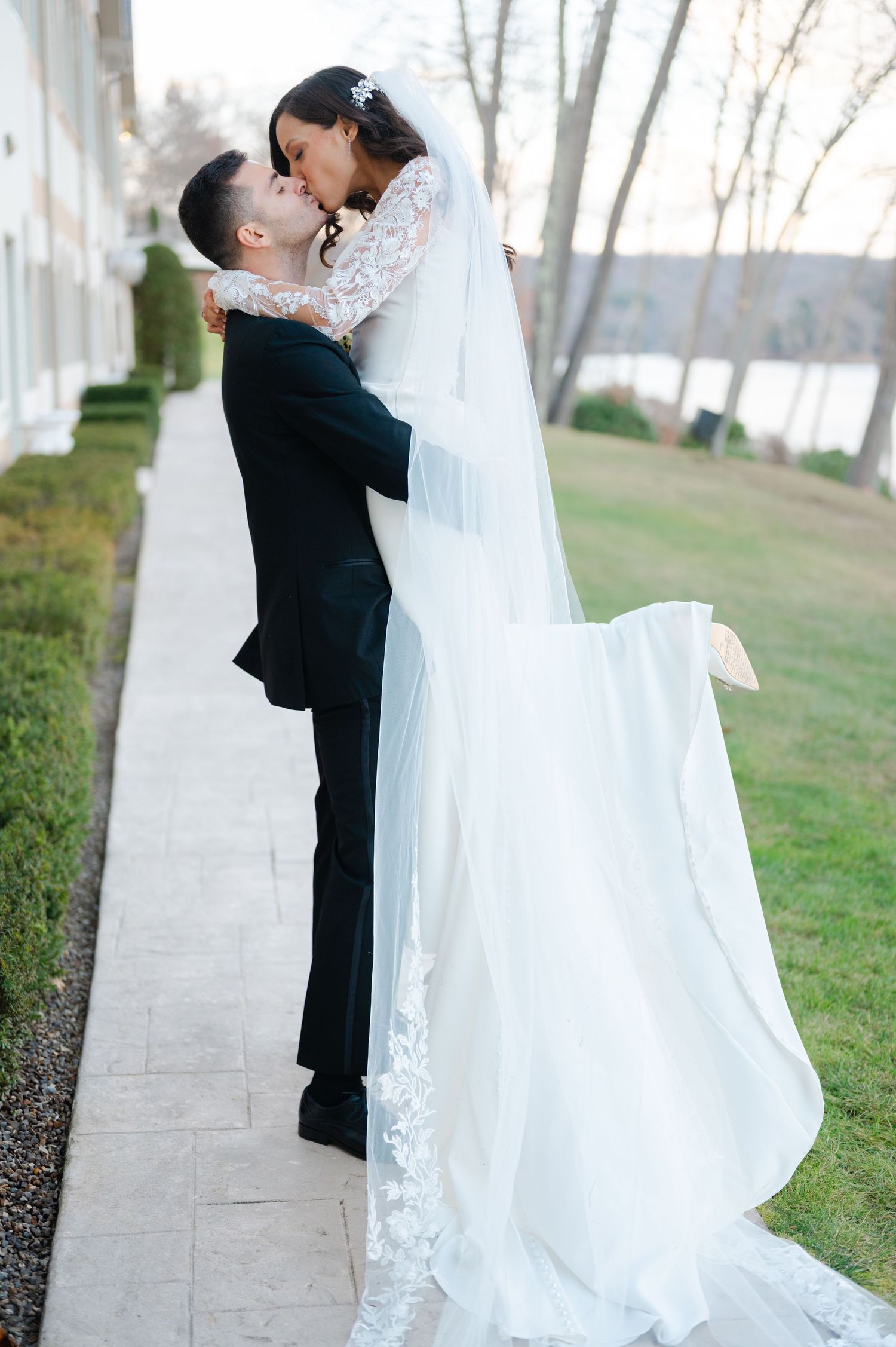 A bride in a flowing white gown and groom in black tuxedo share intimate moments in an outdoor setting with trees.