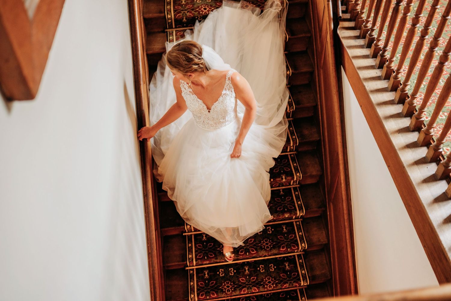 Bride in white wedding dress walking down elegant wooden staircase adorned with ornate railing in sepia tones.