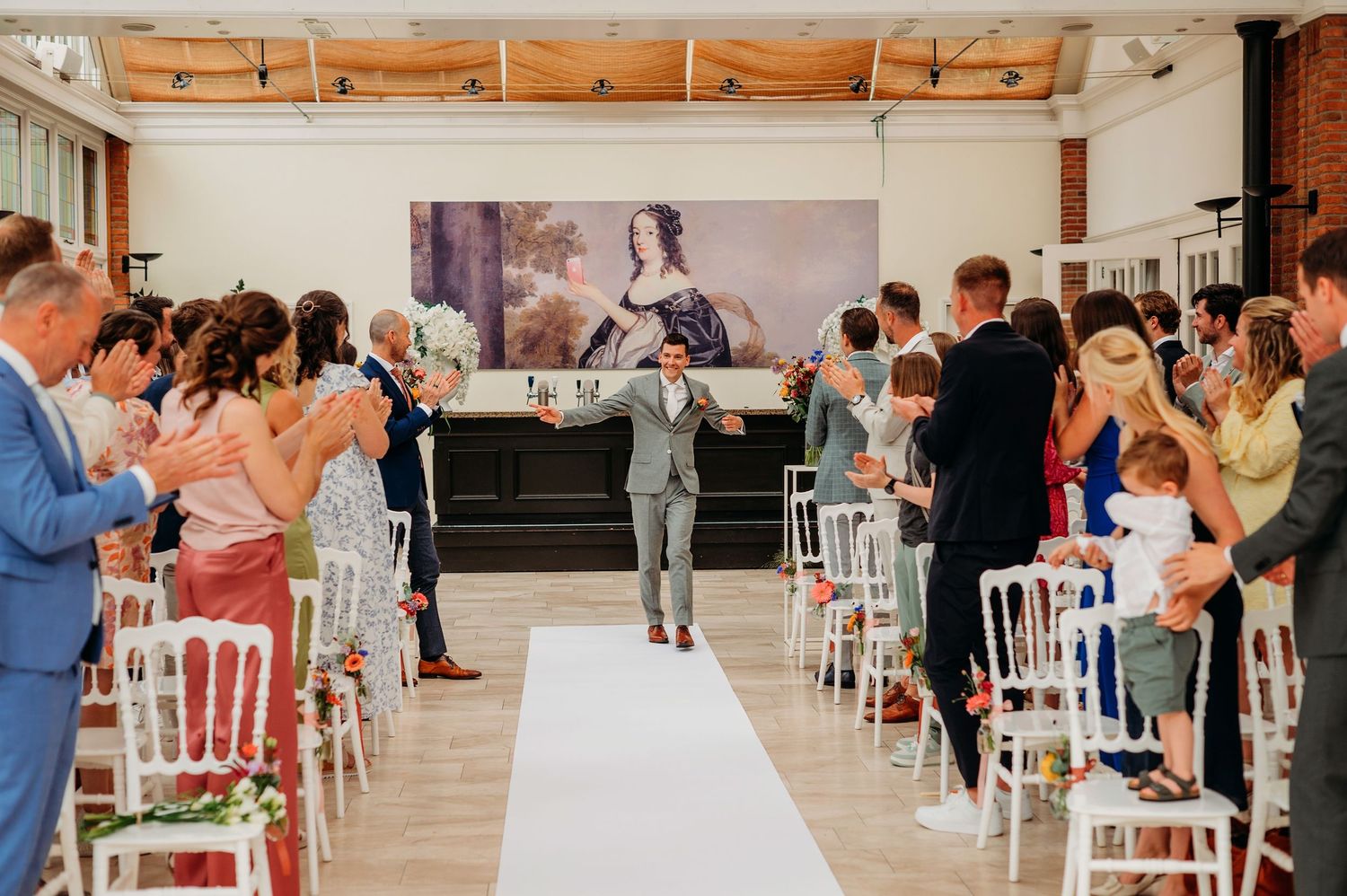 Wedding ceremony guests stand and applaud on both sides of white aisle runner in modern indoor venue.