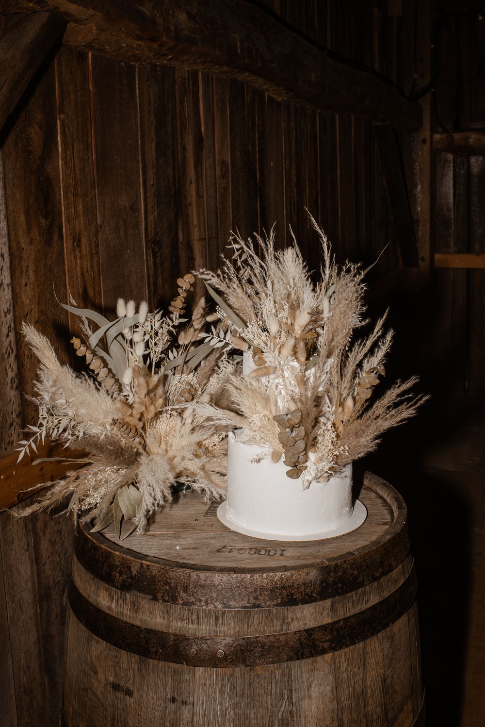White dried pampas grass arrangement in a ceramic vase displayed on a rustic wooden barrel in a barn setting.