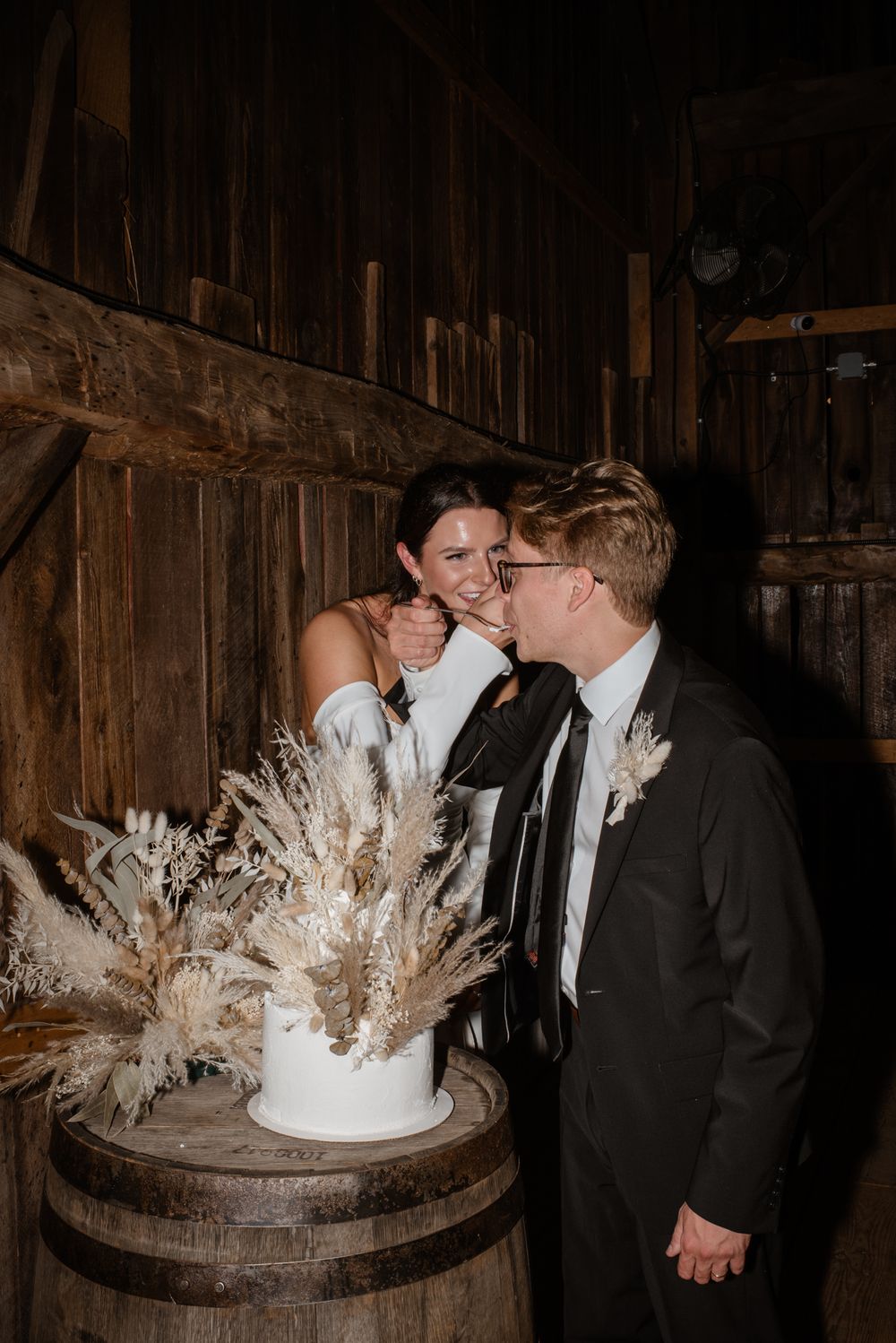 Wedding couple cuts into elegant white cake with dried pampas grass decor atop rustic wooden barrel in barn setting.