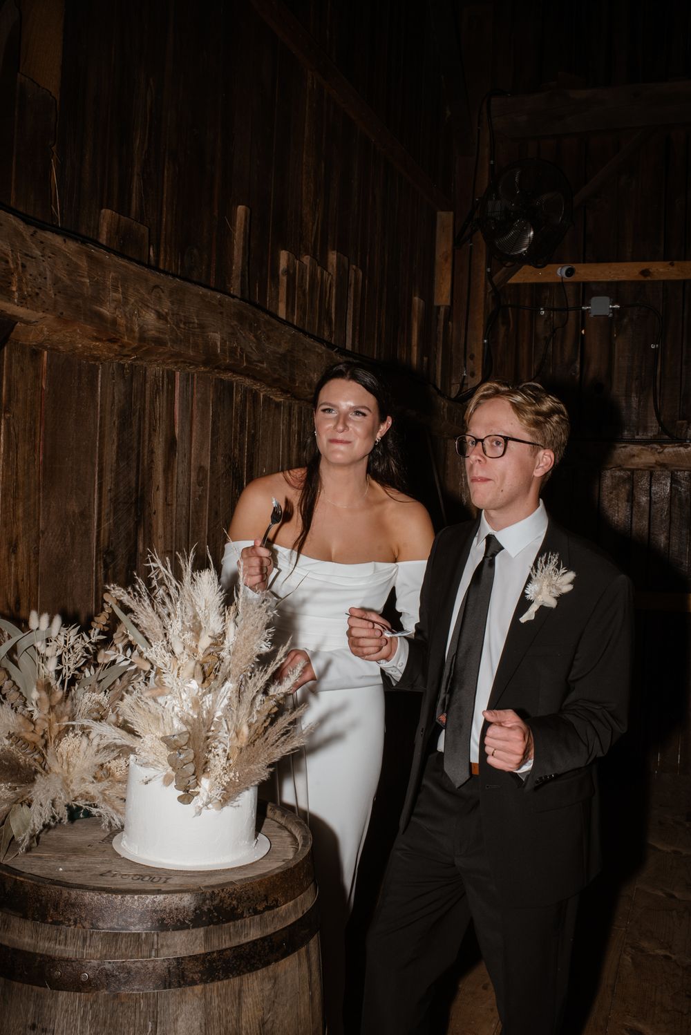 A couple celebrates by their rustic wedding cake decorated with dried flowers set atop a wooden barrel in a barn setting.