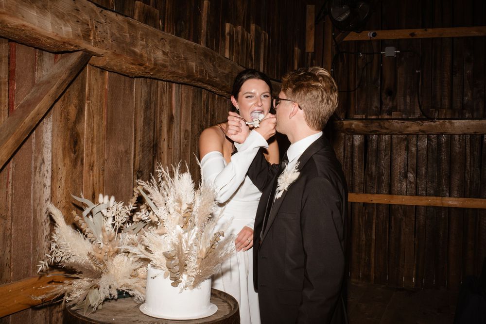 A couple in wedding attire feeds each other cake in a rustic barn setting decorated with pampas grass.