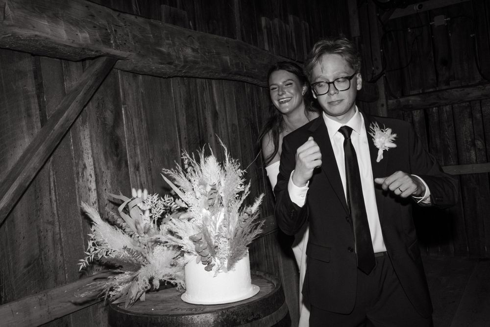 A black and white photo of a couple at a barn wedding celebrating near a floral arrangement decorated cake.
