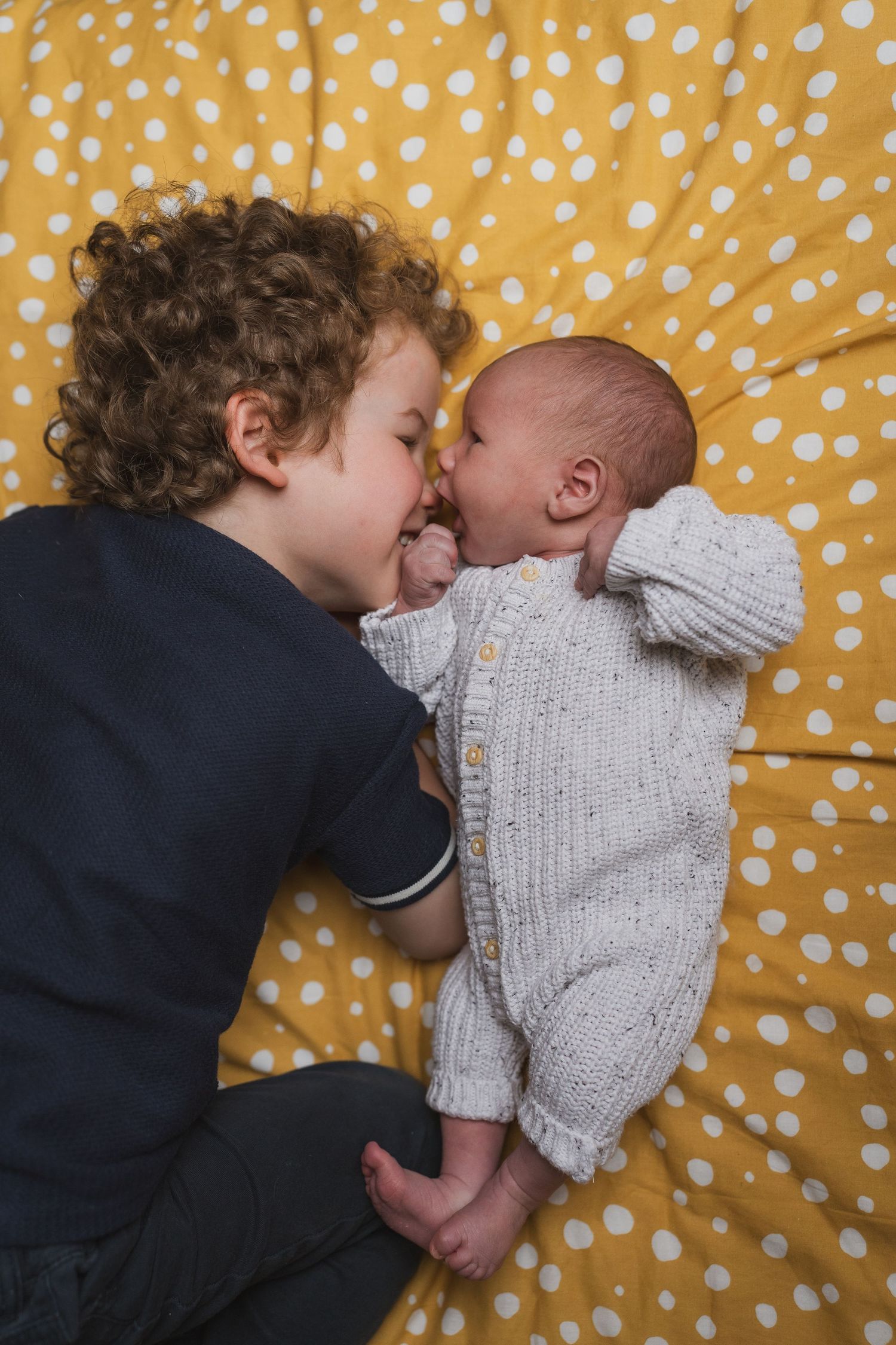Intimate moment between siblings sharing a sweet interaction on a yellow polka dot background during newborn session.