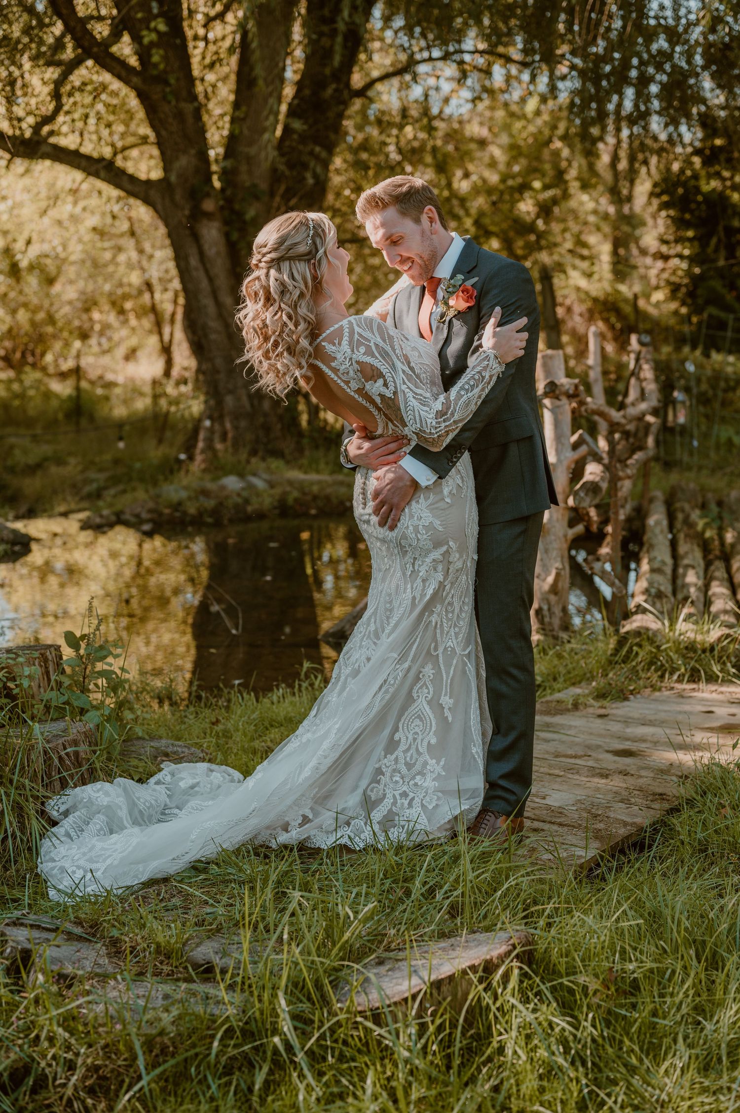 A romantic wedding couple embraces by a tranquil pond at sunset, with the bride wearing an intricate lace gown. Samantha Campbell imagery