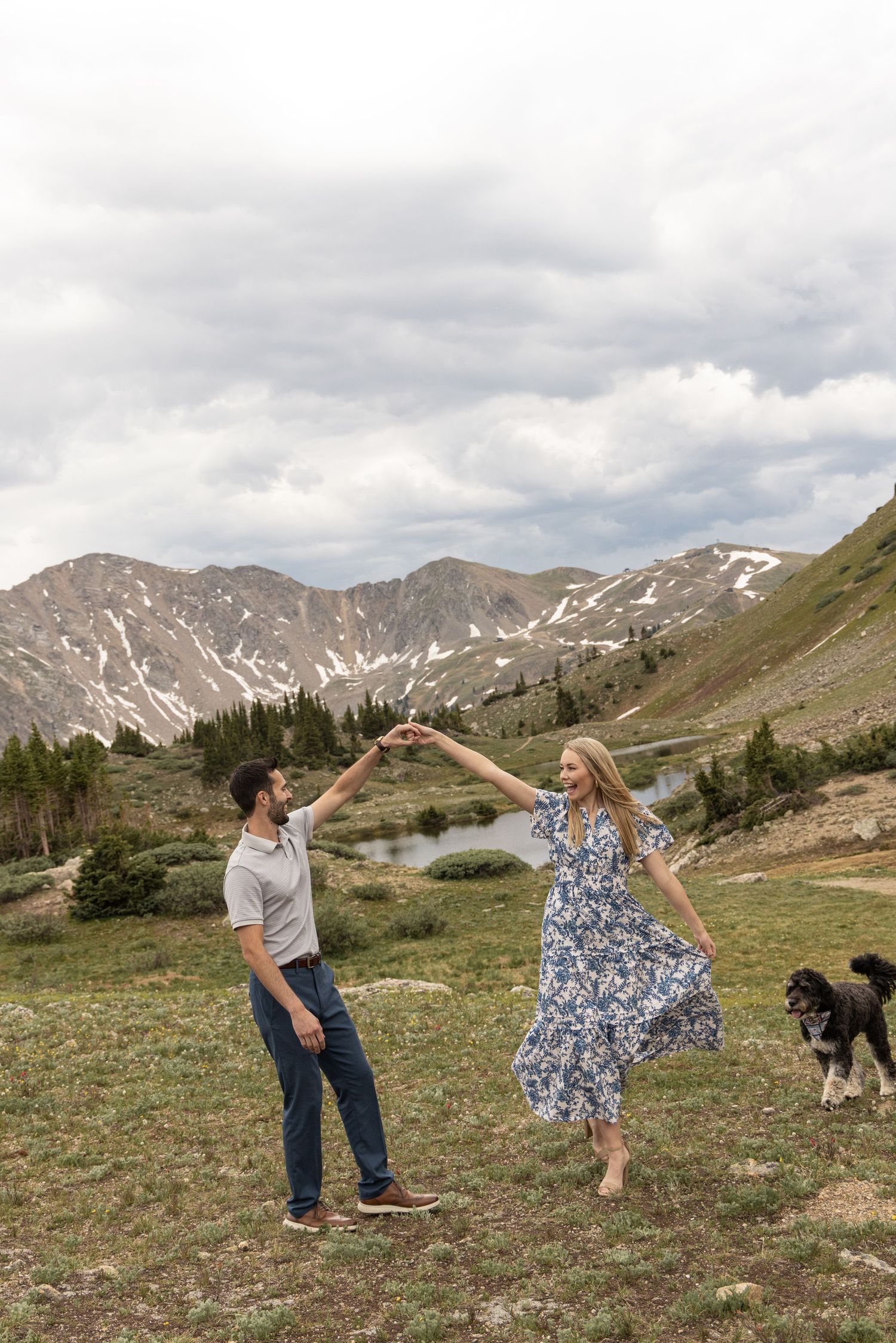 A couple poses playfully with their dog in a scenic mountain meadow with snow-capped peaks in the background.