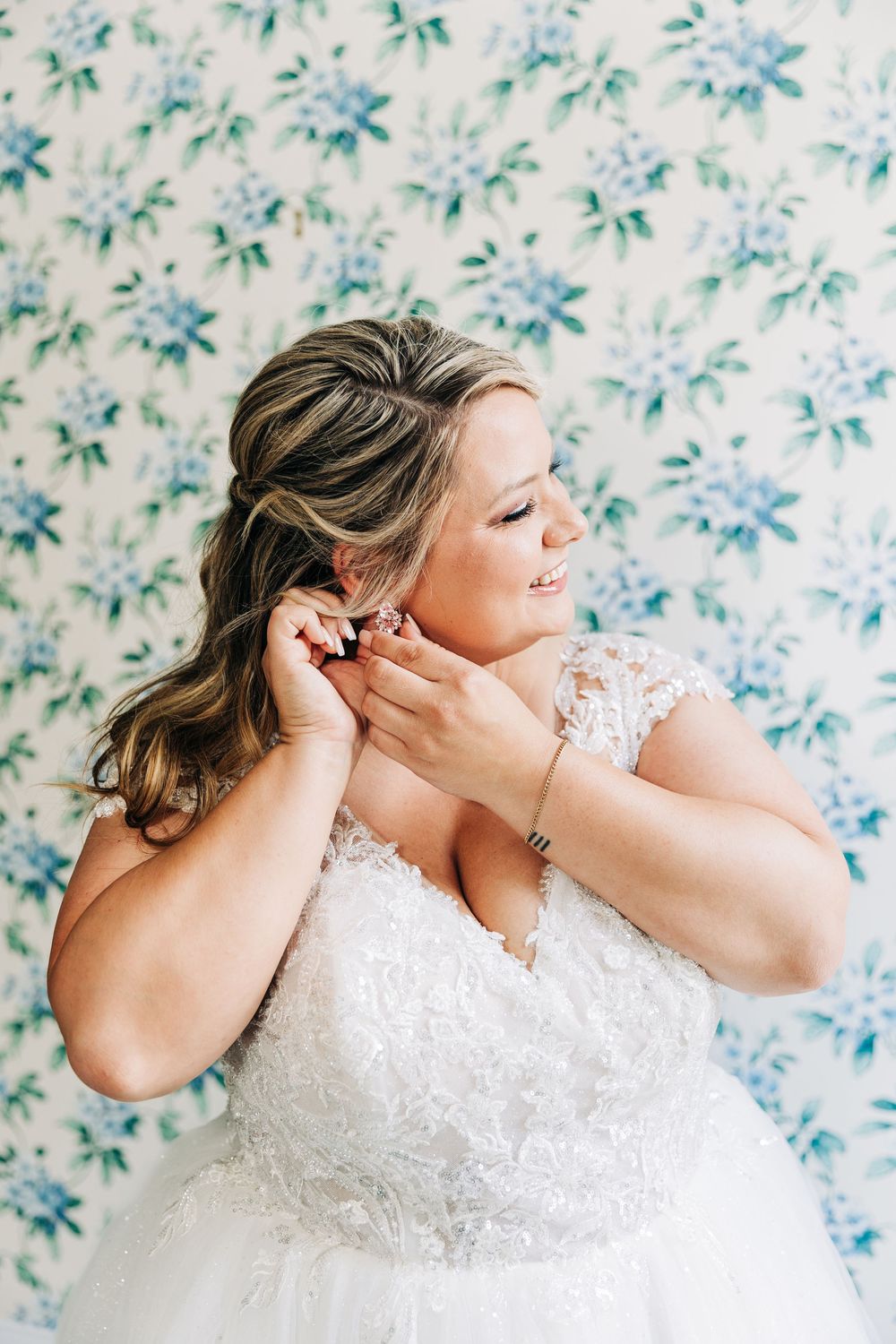 Bride in lace wedding dress puts on earrings against floral wallpaper background.