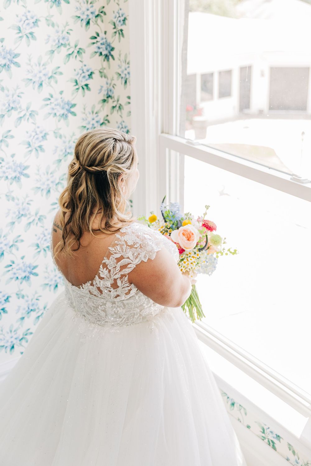 A bride in a white lace gown with a high neckline looks through a sunny window with floral wallpaper.