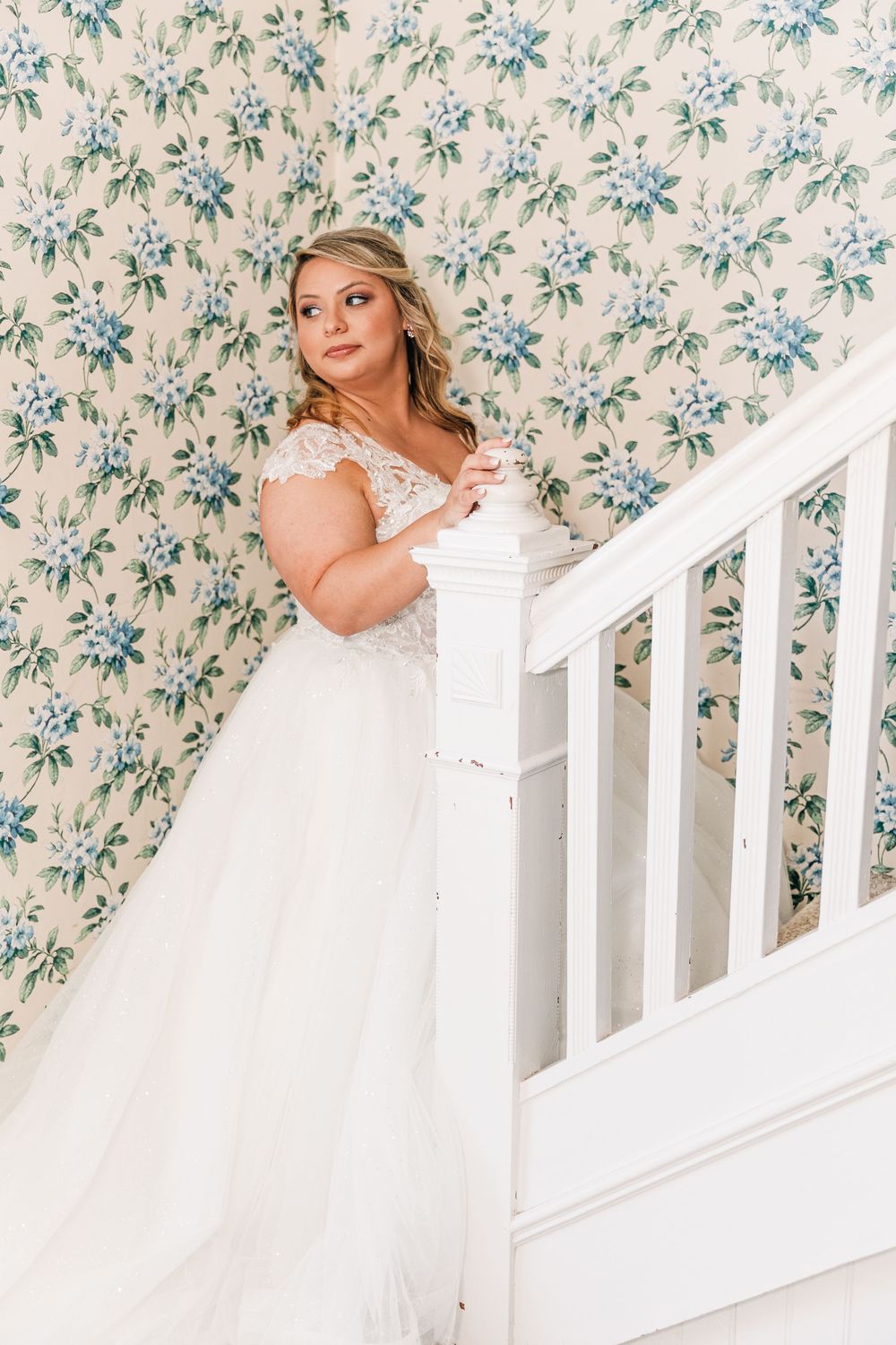 A bride in a white lace wedding gown poses elegantly on a staircase with floral wallpaper background.
