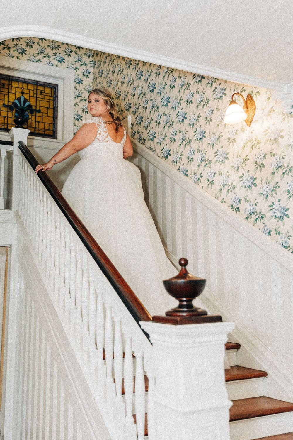 White wedding gown with lace details shown on classic wooden staircase with floral wallpaper and decorative handrail.