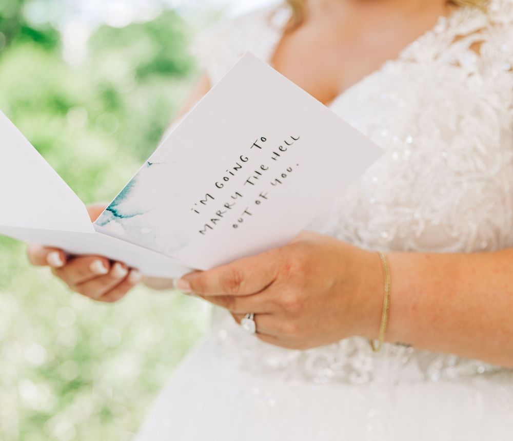 Hands in white lace hold elegant cream-colored wedding invitation cards against a soft blurred green garden background.